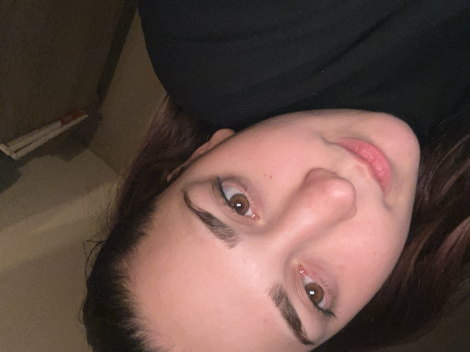 A woman with dark brown hair, fair skin, and brown eyes looks directly at the camera. Her face is slightly tilted. She has well-defined eyebrows and appears to be wearing eyeliner. Her lips are pink. She is wearing a dark-colored garment around her neck. A section of a wall with a white object is visible in the background. The lighting is somewhat dim, casting shadows across the image.