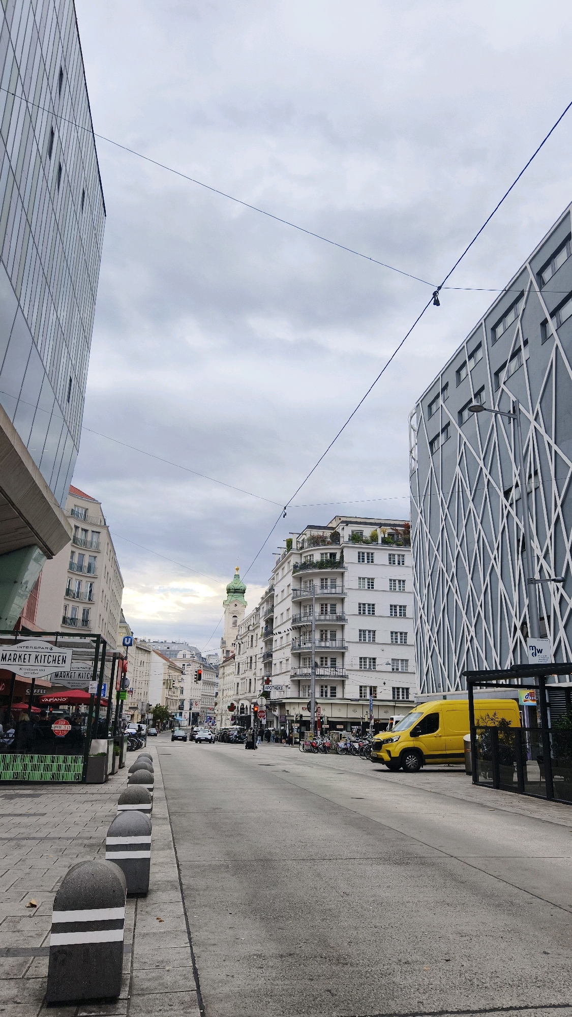 The image depicts a street scene in Vienna, Austria, under a cloudy sky. The perspective is from a sidewalk looking down a moderately wide, paved street lined with buildings on both sides. On the left, a modern glass-fronted building stands tall, with its reflection visible in the glass. Adjacent to it, a building with multiple balconies is visible. Further down the street, a church steeple with a green spire rises above the other structures. On the right side of the street, a contemporary building with a distinctive gray facade featuring a white criss-cross pattern is prominent. In front of this building, a vibrant yellow van is parked. The street is moderately busy with parked motorcycles, cars, and a few pedestrians can be seen. Bollards with reflective white bands line the sidewalk on the left. Signs for "MARKET KITCHEN" and "APOTHEKE" are visible. The overall impression is an urban environment with a mix of architectural styles.