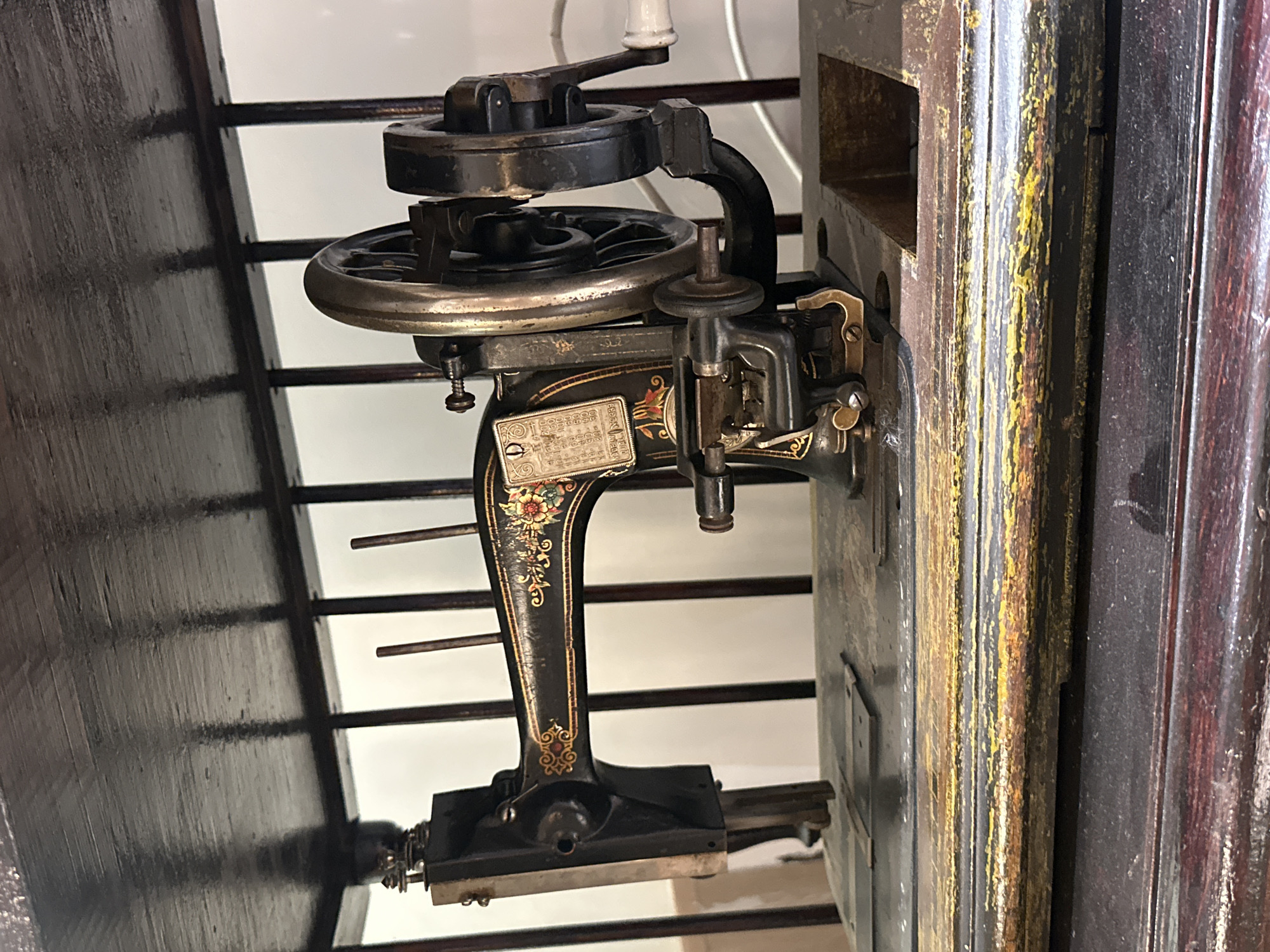An antique black sewing machine, adorned with gold floral patterns, sits on a wooden surface. The machine has a prominent handwheel on its right side, and a metal plate with numbers and lettering is attached to its left. The sewing machine is positioned on a wooden base, which shows signs of wear with chipped paint. Behind the machine, vertical wooden bars are visible, suggesting it is part of a larger piece of furniture or display. The background is predominantly white, with some diffused lighting. This scene is located in the city of Blagoveshchensk, Russia.