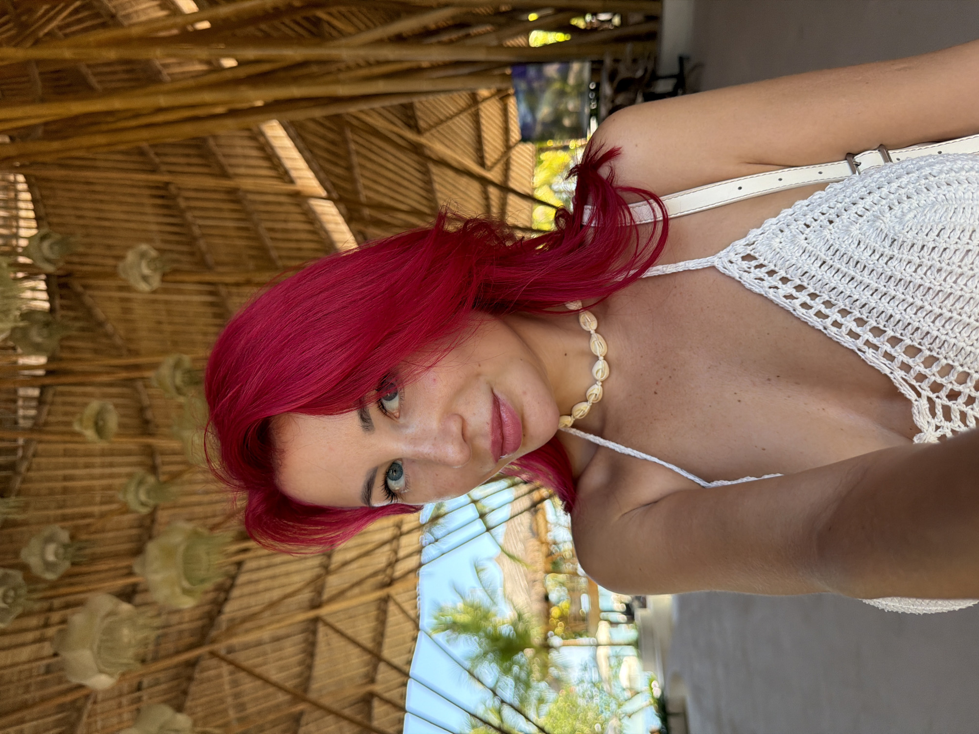A woman with vibrant pink hair, blue eyes, and freckles is taking a selfie in an open-air structure with a thatched roof and bamboo supports. She is wearing a white crocheted bikini top and a shell necklace. The background shows a glimpse of tropical foliage, palm trees, and a body of water, suggesting a beach or resort setting. The overall ambiance is casual and vacation-like. The scene is likely in Kuta, Indonesia, given the tropical environment and architectural style.