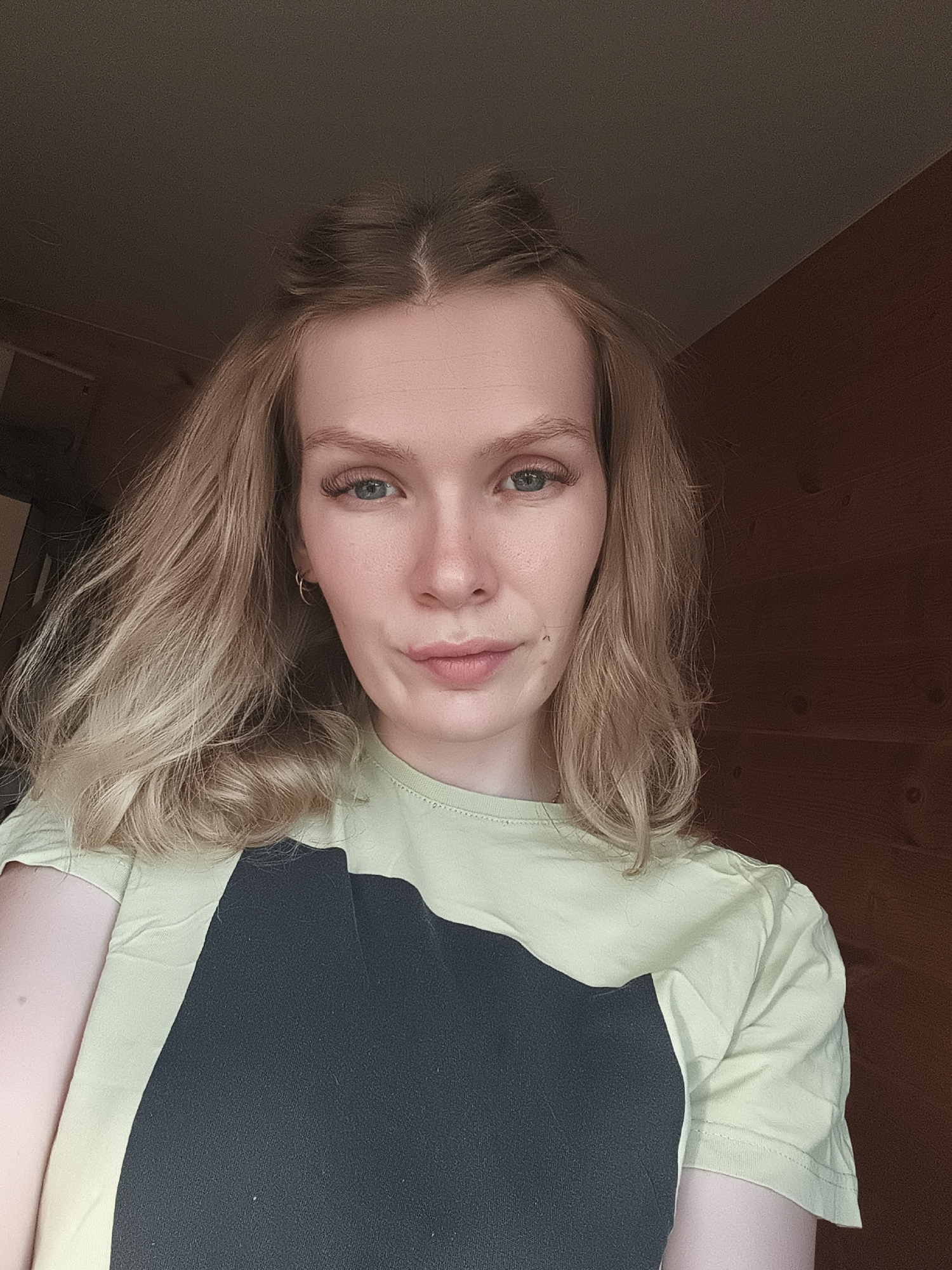 A fair-skinned person with light blonde hair and blue eyes is shown in a selfie. The hair is styled with some strands pulled back from the face. The person is wearing a light green T-shirt with a large, dark-colored geometric design on the front. A small gold hoop earring is visible in one ear. The background appears to be the interior of a room with dark brown wooden wall panels. The exact location in the city of Русский Вожой, Russia cannot be determined from the image.