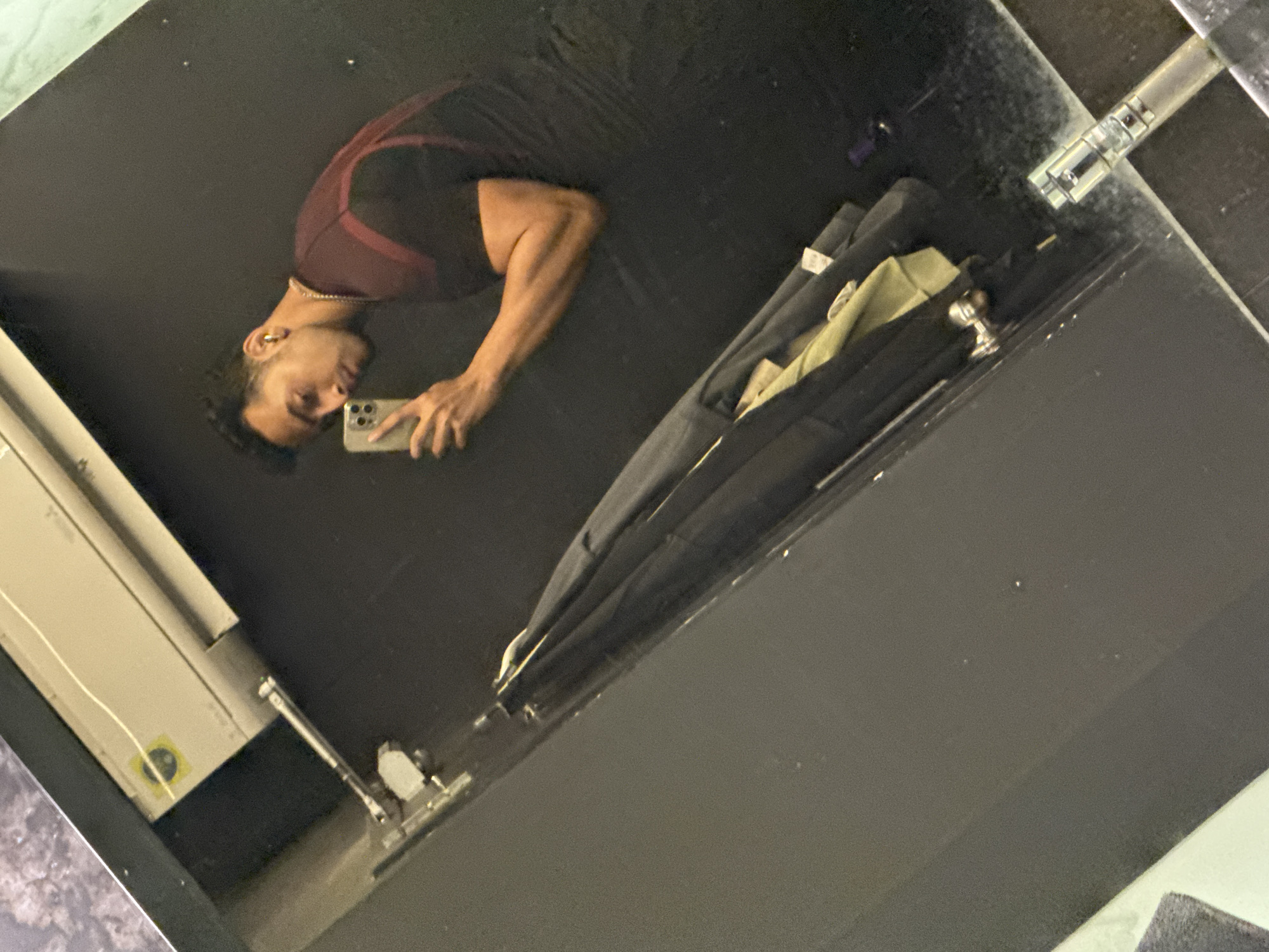 A mirrored image shows a man in a maroon and black athletic top taking a photo of himself with a smartphone. He stands in front of a large, dark reflective surface. To the right of the reflection are a set of what appear to be suit carriers. Behind him, to the left, there is an electrical box. The location or setting within the city of Kolkata, India cannot be determined from the image.