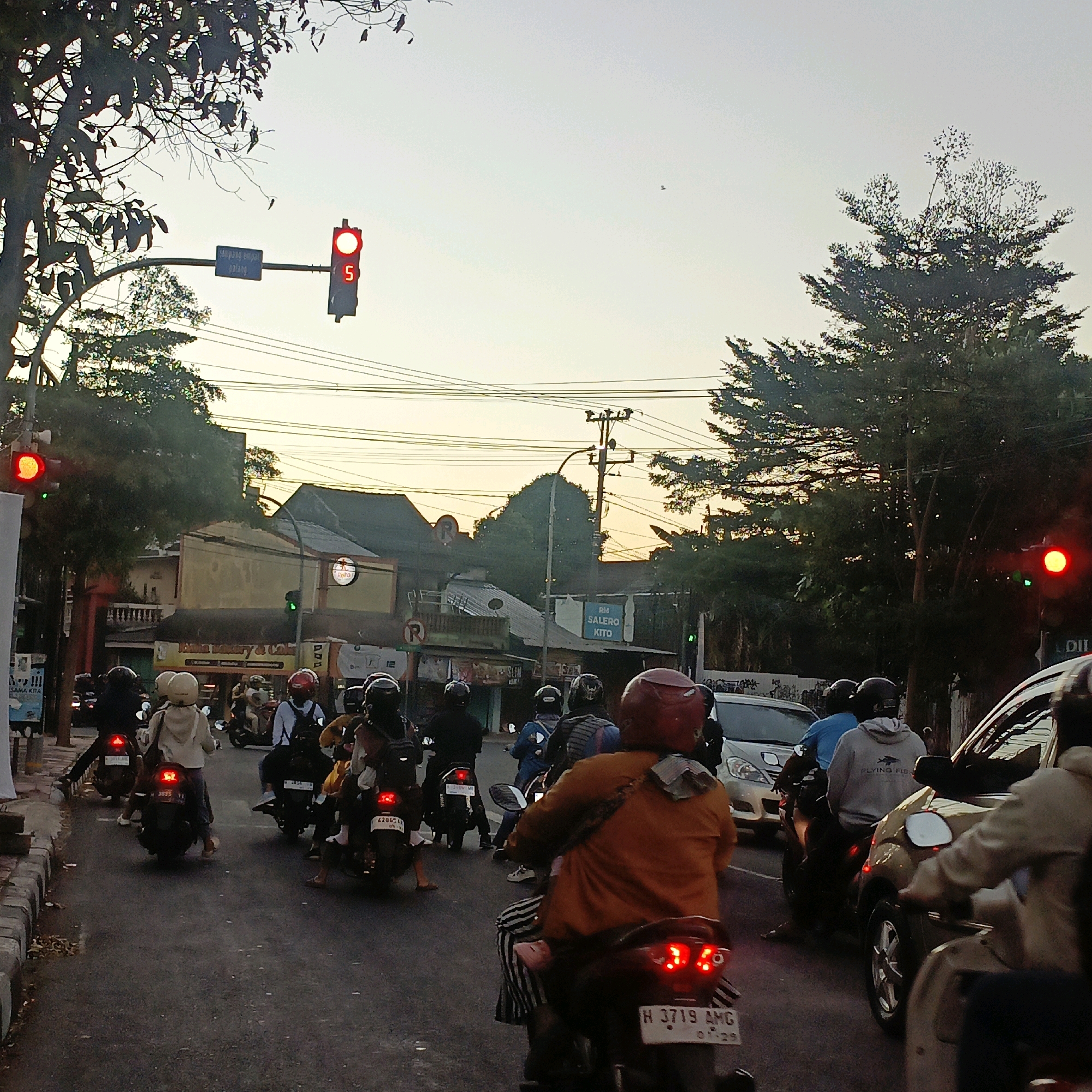 The image depicts a street scene in Salatiga, Indonesia, during what appears to be late afternoon or early evening, with a traffic light indicating red, showing a countdown timer of "5". Numerous motorbikes and a few cars are present on the asphalt road. Most of the people are riding motorbikes, many wearing helmets, and some are carrying backpacks or bags. Buildings line the sides of the street, with trees and power lines overhead. The sky is a pale orange and yellow hue, suggesting sunset. The atmosphere is that of a busy intersection with traffic at a standstill.