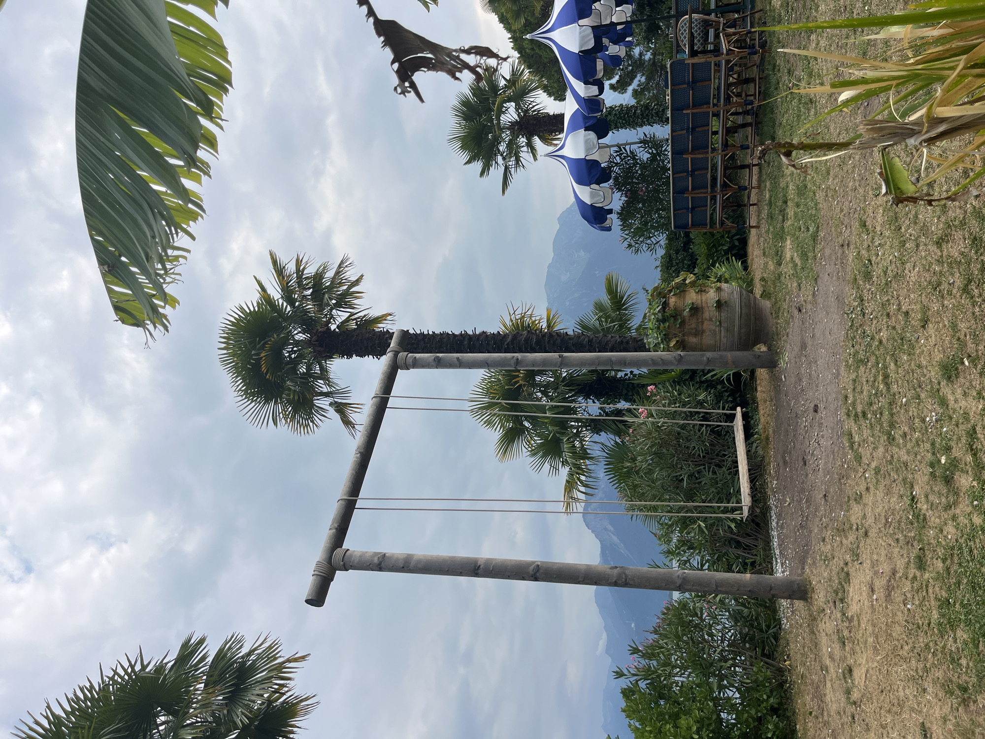 The image depicts an outdoor seating area with a swing set and a view of mountains and a body of water, likely Lake Geneva, in Montreux, Switzerland. In the foreground, a rustic wooden swing set with a wooden seat hangs from thick ropes attached to a horizontal beam supported by two vertical posts. The ground around the swing is a mix of dry grass and dirt. To the right, several large blue and white striped umbrellas provide shade over a seating area with dark-colored chairs and tables. Behind the umbrellas, palm trees and lush green foliage are visible. In the background, a range of mountains rises against a cloudy sky. The scene appears to be a tranquil spot for relaxation, possibly at a restaurant or hotel.