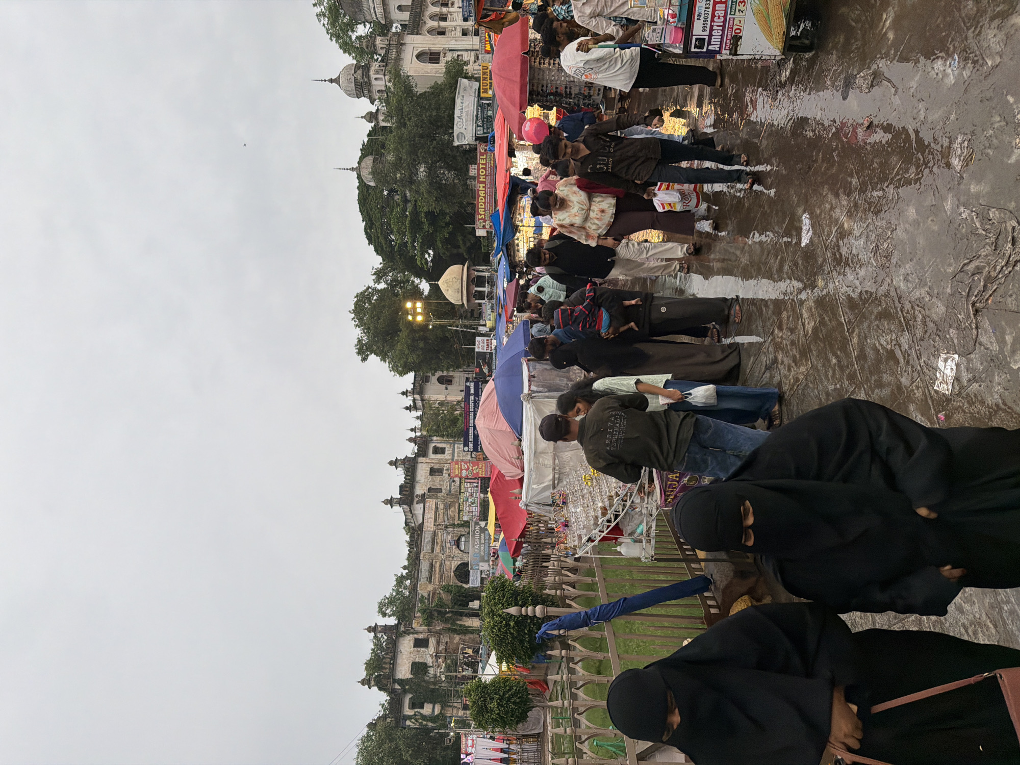The image captures a bustling street scene in Hyderabad, India, likely near Charminar, given the architectural style of the buildings in the background which feature domes and minarets. The foreground is dominated by a crowded marketplace with numerous stalls and people. A significant portion of the pavement is wet and reflects the surrounding lights and figures, suggesting recent rain or a wet environment.
Several individuals are visible, many of whom are dressed in traditional Indian attire. Two women in the immediate foreground are wearing black burqas that cover their faces, with one carrying a pink handbag. Other people in the scene are dressed in a variety of clothing, including casual shirts, t-shirts, and trousers, with some women in kurtis and headscarves.
The marketplace features multiple colorful umbrellas and canopies over various stalls selling goods. Items visible on display include what appear to be jewelry or trinkets on a table, and possibly food items on a cart in the right foreground. Signage for "SADDAM HOTEL" and "American Corner" are visible, indicating commercial establishments. The crowd is densely packed, with people walking in various directions, browsing the stalls, and interacting with each other. The overall atmosphere appears to be one of vibrant street commerce under an overcast sky. The architectural style of the buildings, with their ornate facades and domed structures, strongly suggests a historical and culturally significant area of Hyderabad.