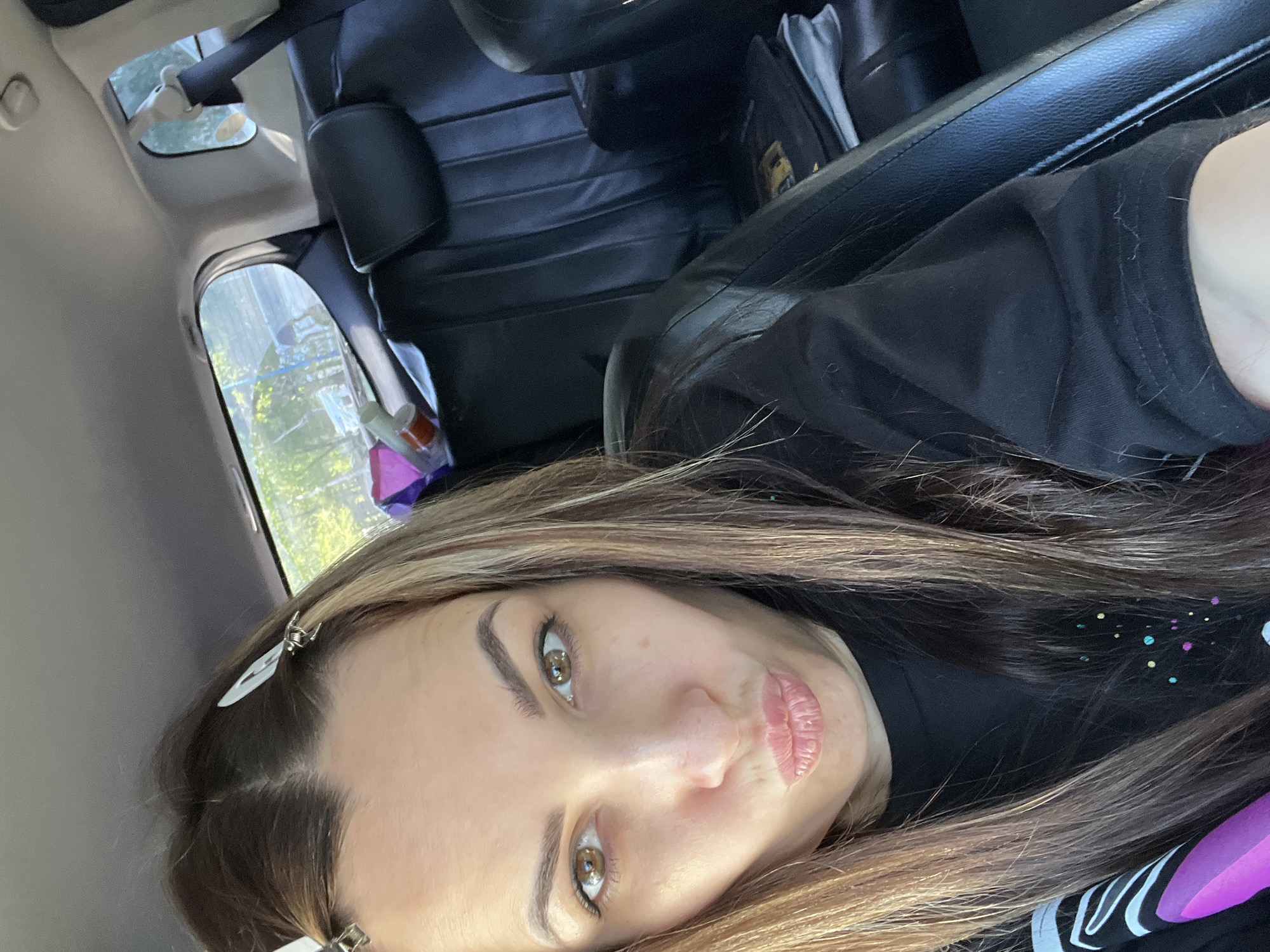A selfie taken inside a car shows a woman with light brown hair and light eyes, looking directly at the camera with her lips pursed. She is wearing a black shirt with colorful speckles. The background reveals the car's interior, including black leather seats, the dashboard, and a glimpse of the outside through the car window. The location is unknown, but visible greenery outside the window suggests an urban or suburban environment.