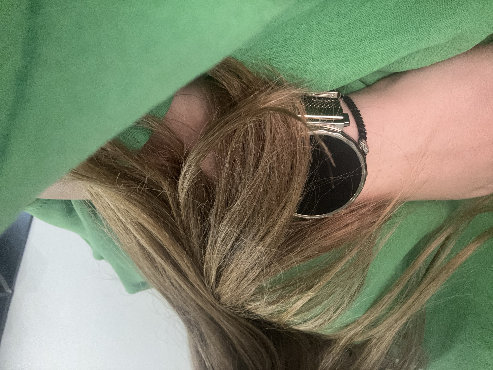 The image is a close-up, high-angle shot focusing on a person's arm, wrist, and hair. The person is wearing a green garment, possibly a hoodie or sweatshirt, which dominates the background. On the wrist, a silver smartwatch with a black face is clearly visible. The person's hair, a mix of blonde and brown tones, is spread across the frame, partially obscuring their arm and the watch. Some strands of hair appear to have a subtle green tint, possibly from dye or interaction with the garment. The lighting suggests an indoor setting, possibly during the daytime, with no indication of specific weather conditions or a precise location within Burgas, Bulgaria. The overall impression is intimate and personal, with the focus on the texture of hair and fabric, and the detail of the watch. There is no visible text in the image.