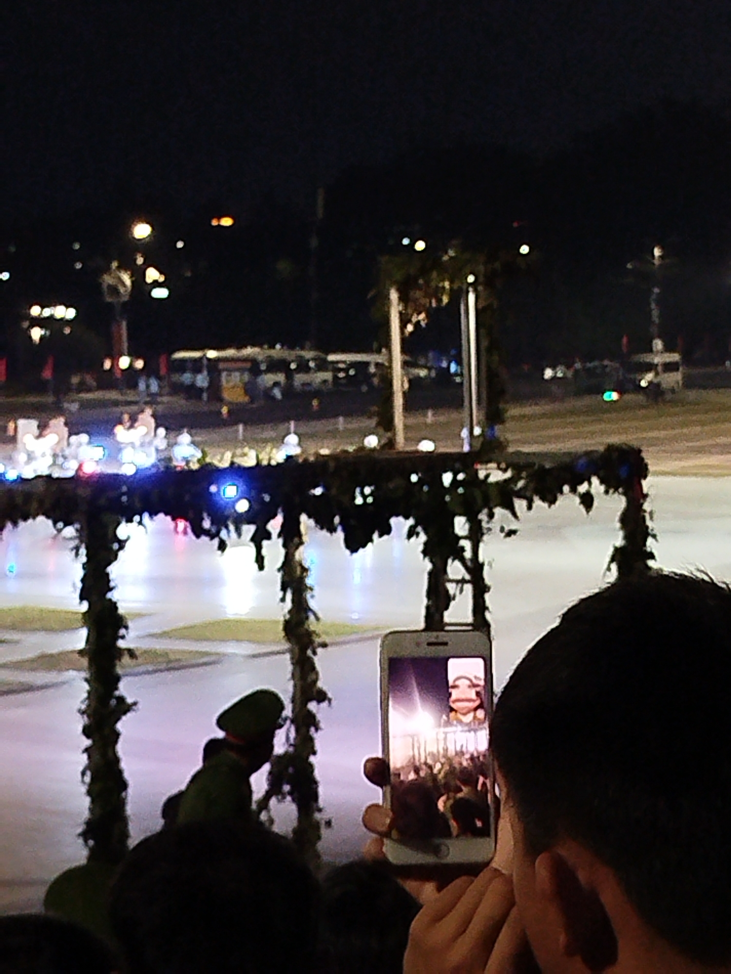In Da Lat, Vietnam, a nighttime event is captured, possibly a parade or public gathering, with a person holding up a smartphone to record the scene. The phone screen displays a live video feed showing a person in a suit and a crowd in the background. In the foreground, another person, wearing a dark green uniform and hat, is visible from the side, facing away from the camera, potentially participating in or observing the event. The background is illuminated by various lights, including streetlights and lights from vehicles, suggesting an urban setting. The overall atmosphere is dark, with artificial lighting creating a festive or ceremonial mood.