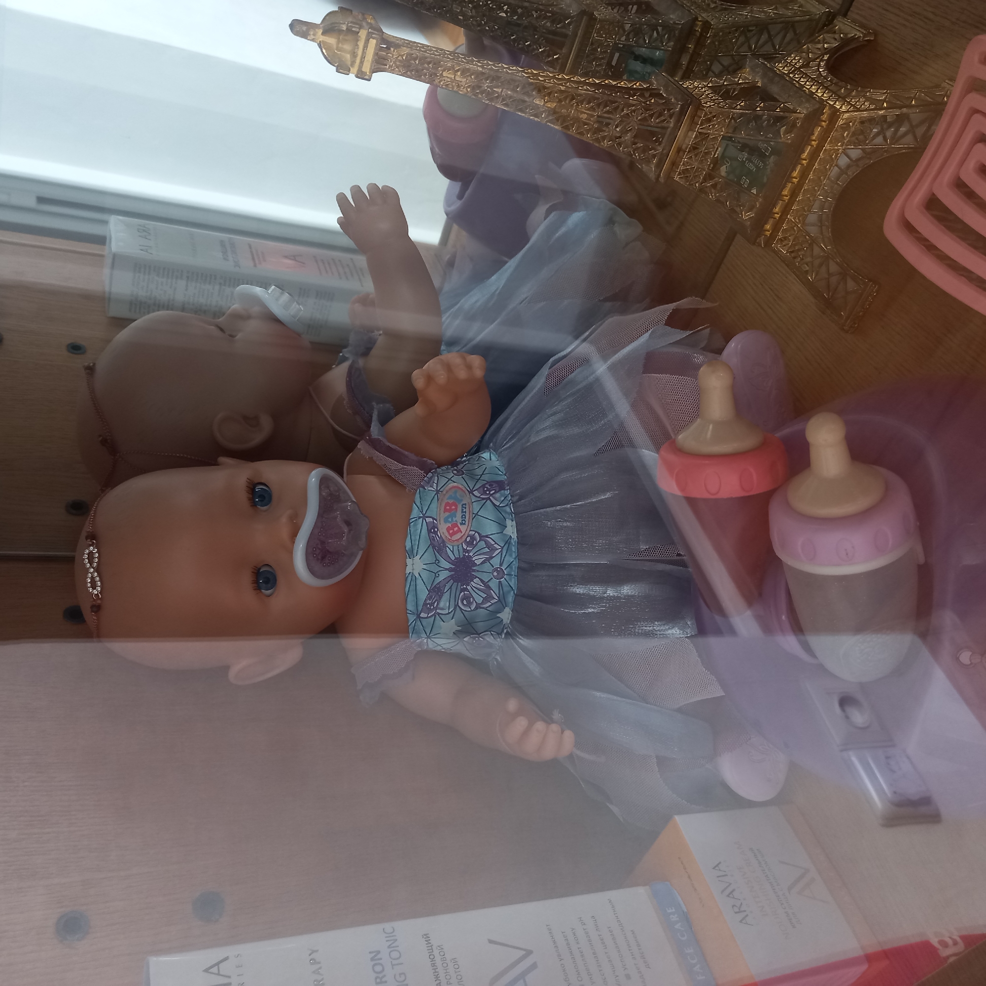 The image shows a baby doll wearing a blue and silver dress with butterfly motifs and a pacifier. The doll is positioned inside a display case, reflected in the glass. To the left of the doll, there are cosmetic product boxes. To the right, a miniature golden Eiffel Tower replica is visible. In the foreground, two baby bottles are present. The setting appears to be a shop or a home display in Cepeleuți, Moldova. The lighting is dim, and the image is slightly blurry due to reflections.
