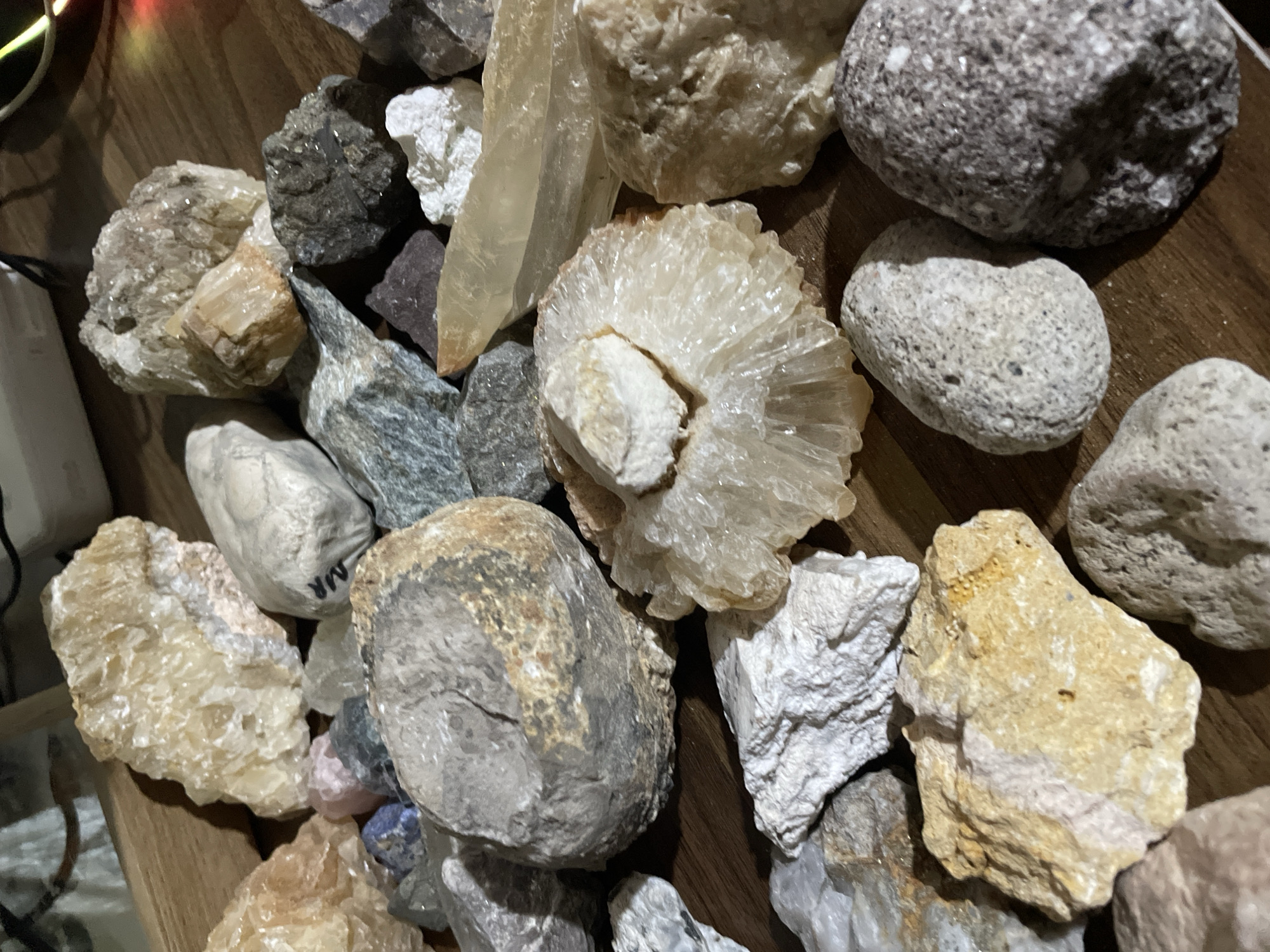 The image displays a close-up, high-angle view of a diverse collection of rocks and mineral specimens scattered across a warm brown wooden surface, likely a table or shelf. No people are visible.
The collection features a variety of shapes, sizes, and textures. Prominently in the center-right is a large, light-colored crystalline formation with a radiating, fan-like or rosette structure, possibly a type of calcite or quartz druzy. Several other translucent to opaque white, beige, and light brown crystals and raw mineral pieces are distributed throughout the pile, some showing distinct facets and cleavage planes. Among these are a few larger, irregularly shaped grey and dark grey rocks, some appearing porous or volcanic, and others more rounded like river stones. One dark grey rock near the top-left has visible shiny flecks.
A long, translucent, light amber-colored crystal extends from the top-center downwards. Smaller, subtly colored stones, including hints of purple, pink, and blue, are also nestled among the larger pieces.
In the top-left corner, an out-of-focus light source casts a soft, colored glow (red and yellow), and a white cable or object is partially visible. In one of the cropped views, a white object with the letters "MR" written in black is discernible, possibly a label or tag for one of the specimens.
The scene is an indoor, still-life arrangement, suggesting a personal collection or display. No specific activity is taking place other than the static presentation of the minerals. The lighting indicates an indoor setting, likely under artificial light.