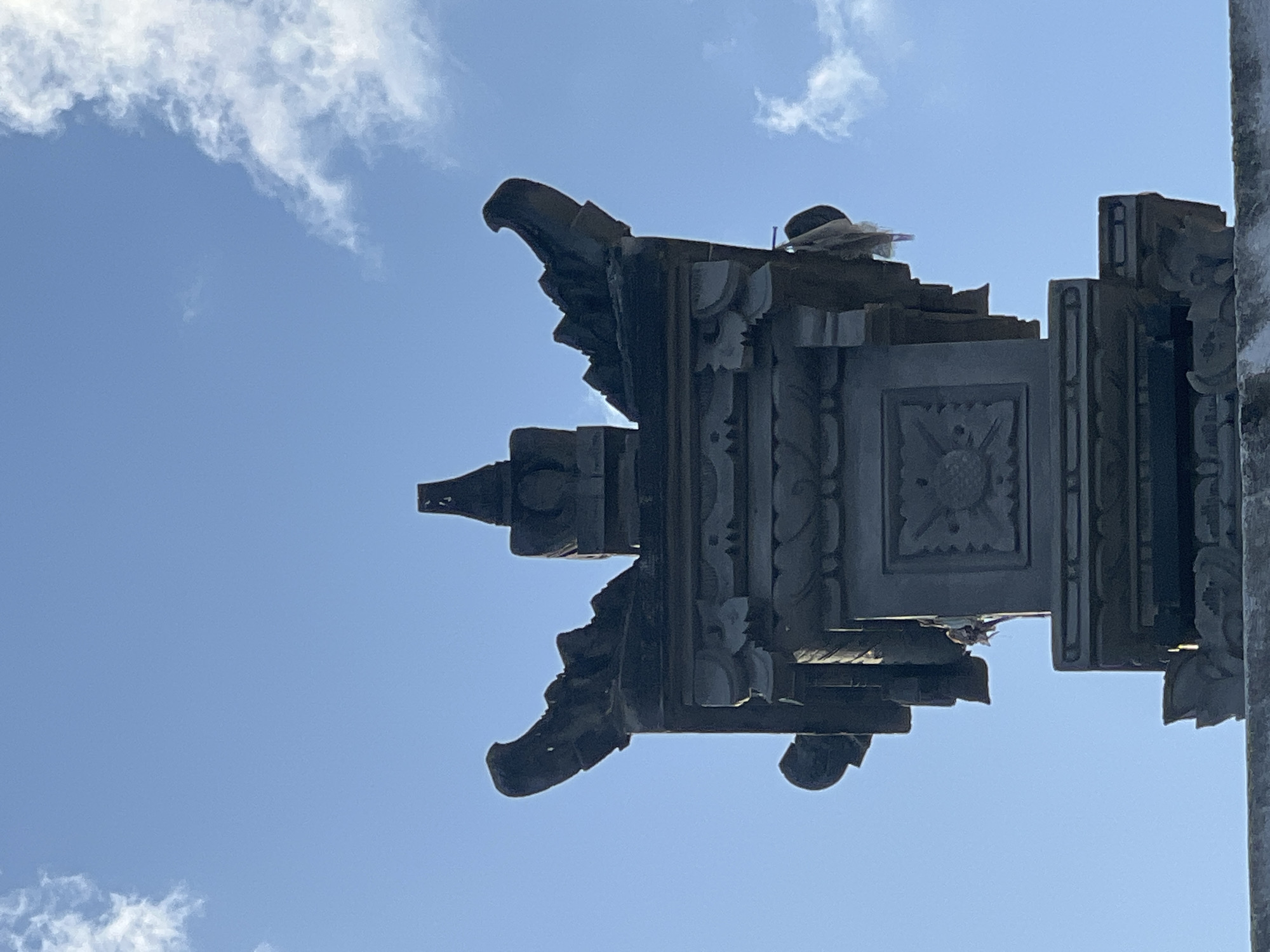 The image shows a close-up, low-angle view of an ornate stone structure, likely a temple or shrine ornament, against a bright blue sky with scattered white clouds. The structure is dark gray stone and features intricate carvings, including decorative elements on the roofline that resemble mythical creatures or stylized eaves. A square panel with a floral or sun-like motif is visible on one side of the structure. Small pieces of white cloth are tied to various points on the ornamentation, suggesting they may be prayer flags or offerings. The structure appears to be ancient or made in a traditional style. The environment is an outdoor setting with clear daylight. The architecture suggests a Balinese or Southeast Asian origin, consistent with locations in Denpasar, Indonesia.