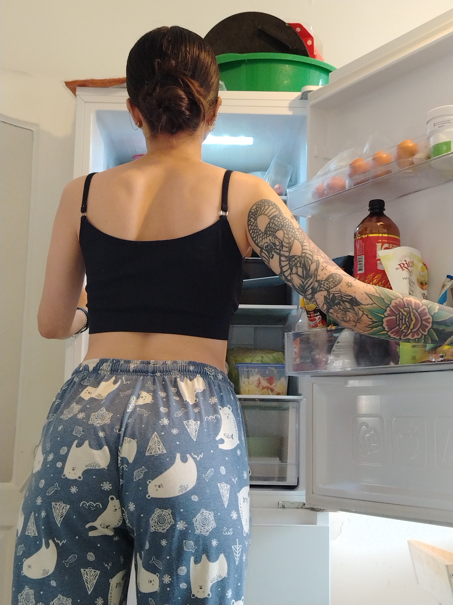 A person stands with their back to the viewer, facing an open white refrigerator in a domestic setting, located in Noviy, Russia. The individual, appearing female, has dark hair styled in a low bun and wears a black sleeveless ribbed crop top with adjustable straps, along with blue pajama pants featuring a white pattern of polar bears, trees, and other shapes. Multiple small hoop earrings are visible in both ears. The person's left arm is extended into the illuminated refrigerator, revealing a full sleeve tattoo comprising a large snake, various floral designs, leaves, and stars. The right arm hangs loosely by their side.
Inside the refrigerator, the door shelves contain a white container and a green object on the top shelf, several oranges or mandarins packaged in a clear plastic bag on the middle shelf, and a dark brown glass bottle alongside a yellow and red package of mayonnaise (Mr. RiCo brand with Cyrillic text) on the bottom shelf. The main compartment shows a clear plastic container with prepared food (possibly salad) on a lower shelf, and the interior door has molded indentations for produce. Above the refrigerator, a wooden shelf is visible, and atop the appliance sits a large green plastic basin with a partially covered black lid, next to a red and white patterned textile. The surrounding walls are white.