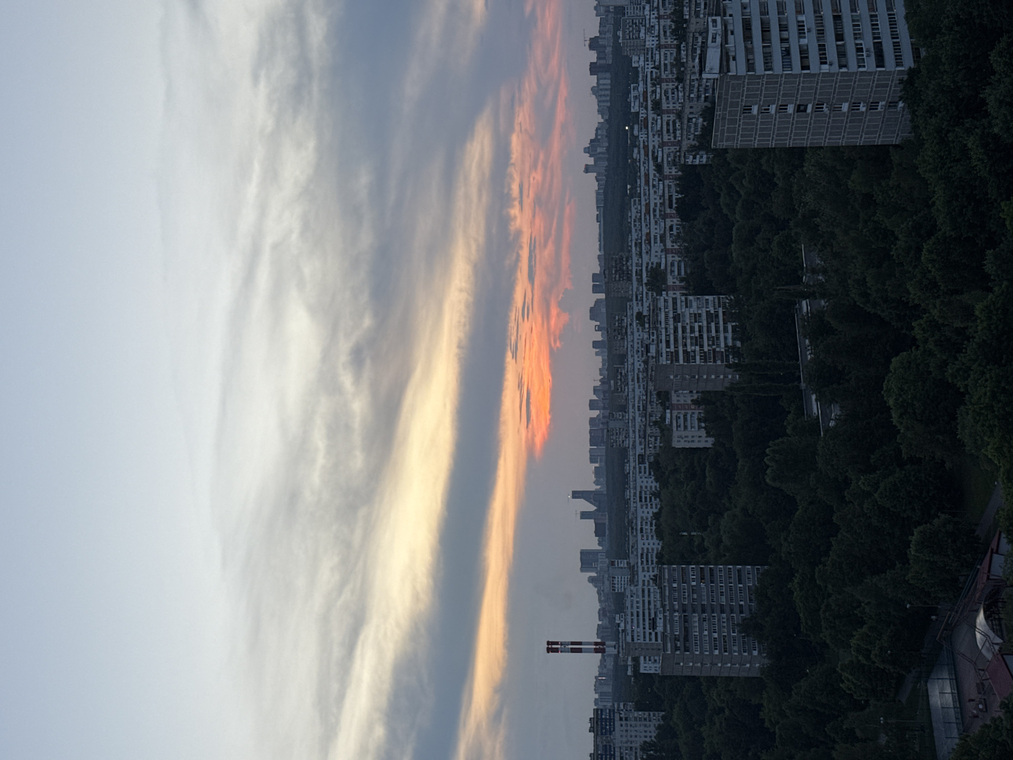 An aerial view captures a panoramic cityscape of Moscow, Russia, at dusk. The sky is a gradient of pale blue at the top, transitioning to soft yellows and oranges from a setting sun, with streaks of grey clouds. Below, a dense forest separates two distinct areas of the city. In the foreground, on the left, a multi-story residential building is partially visible, with a tennis court and a covered structure in front of it. The middle ground is dominated by rows of apartment buildings, interspersed with a significant amount of lush green trees. A prominent industrial chimney with red and white markings stands tall on the left side of the cityscape. In the background, a distant skyline of taller buildings, including skyscrapers, is silhouetted against the twilight sky. The overall scene depicts a residential area within Moscow, characterized by a mix of apartment blocks, extensive greenery, and industrial elements.
