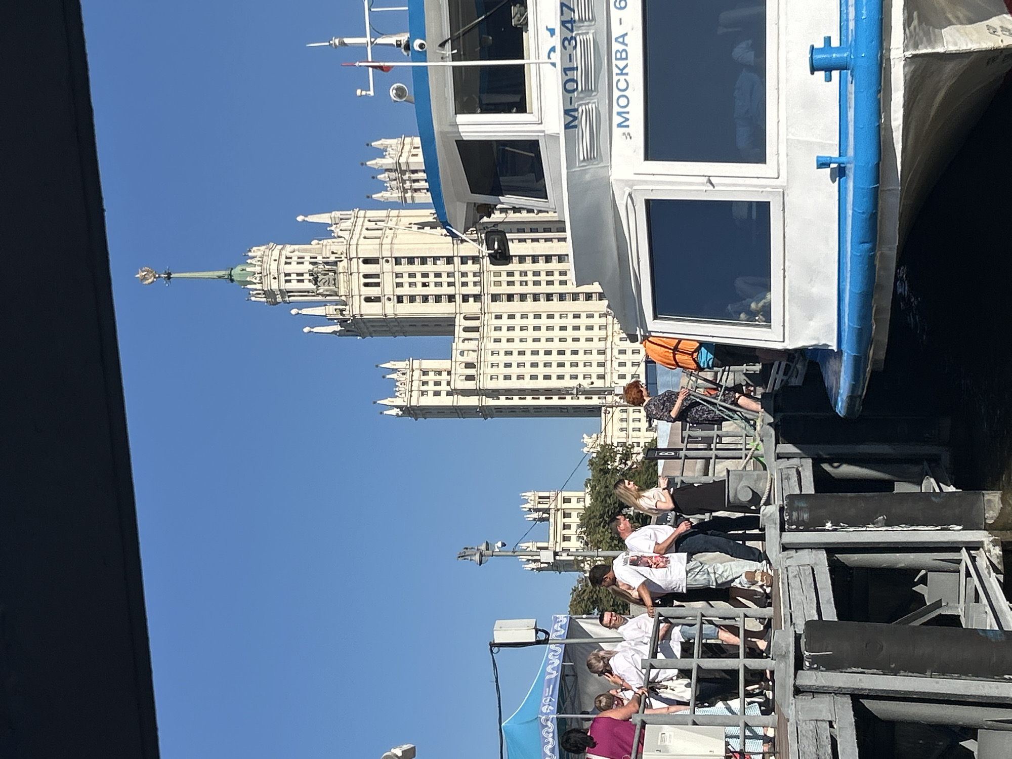 This is a full-shot, eye-level, outdoor, daylight photograph taken on a clear day with a blue sky. In the foreground, a white boat with a blue stripe along its top edge is docked. The boat has large windows and text reading "M-01-347" and "МОСКВА - 6" on its side. A group of people are standing on a metal pier to the left of the boat, some looking towards the water, others engaged in conversation. A woman in a purple top stands out on the far left, while others are dressed in white shirts and casual attire. Behind the people and pier, several imposing Stalinist-era skyscrapers dominate the background, one with a prominent spire topped by a golden eagle. Trees and smaller buildings are visible between the pier and the skyscrapers. The scene is a waterfront area in Moscow, Russia, likely a tourist location given the presence of the boat and the architectural landmarks.