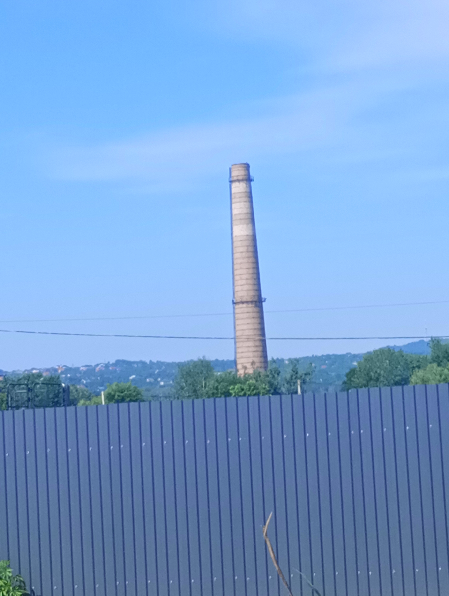 A tall, weathered industrial smokestack stands prominently against a clear blue sky. The smokestack is cylindrical and tapers slightly towards its top, with distinct horizontal bands of differing shades of beige and gray, suggesting age and exposure to the elements. A small, dark structure is visible near its peak.
In the foreground, a dark gray, corrugated metal fence runs horizontally across the frame, obscuring the lower portion of the scene. Behind the fence, a line of lush green trees forms a dense barrier, with a hint of a town or village nestled in the distant rolling hills. The landscape suggests a rural or semi-rural setting. A thin, dark wire is stretched across the sky above the trees, likely a power line.
The overall impression is one of a sunny day, with the bright blue sky indicating fair weather. The time of day appears to be daytime, likely late morning or afternoon, given the brightness and the lack of distinct shadows. There is no text visible in the image. The scene evokes a sense of industrial history contrasting with the natural landscape.