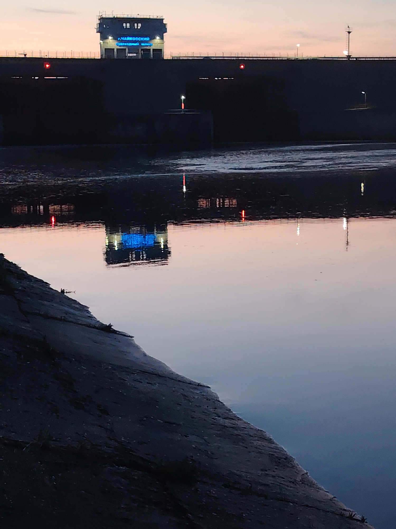 The image depicts the Chaikovsky ship lock in the city of Chaikovsky, Russia at dusk or dawn. The concrete embankment is in the foreground, leading to a body of water reflecting the lock's structure. The lock itself is silhouetted against a colorful sky, with blue lettering visible on the structure. The water surface shows light reflections from the city lights. The scene conveys a sense of stillness and tranquility.