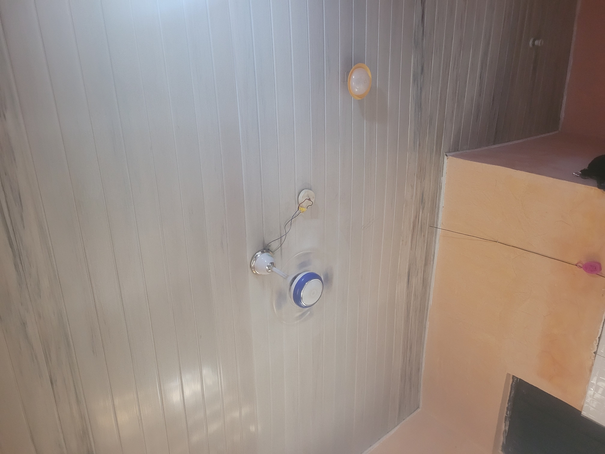The image shows a ceiling with a light fixture and a ceiling fan. The ceiling is made of white plastic panels with vertical grooves. There are two lights visible, one is a circular orange fixture, and the other is a white fixture with a blue and white fan attached. The fan appears to be in motion, as indicated by the blur. To the right of the ceiling, a portion of a wall is visible, painted a peachy orange color. A dark object, possibly a piece of furniture, is partially visible in the bottom right corner. A thin black wire extends from the ceiling fan across the image towards the orange wall. A small pink object, possibly a decoration, is attached to the orange wall near the top right. The scene appears to be indoors, likely in a residential or commercial space. There is no text visible in the image.