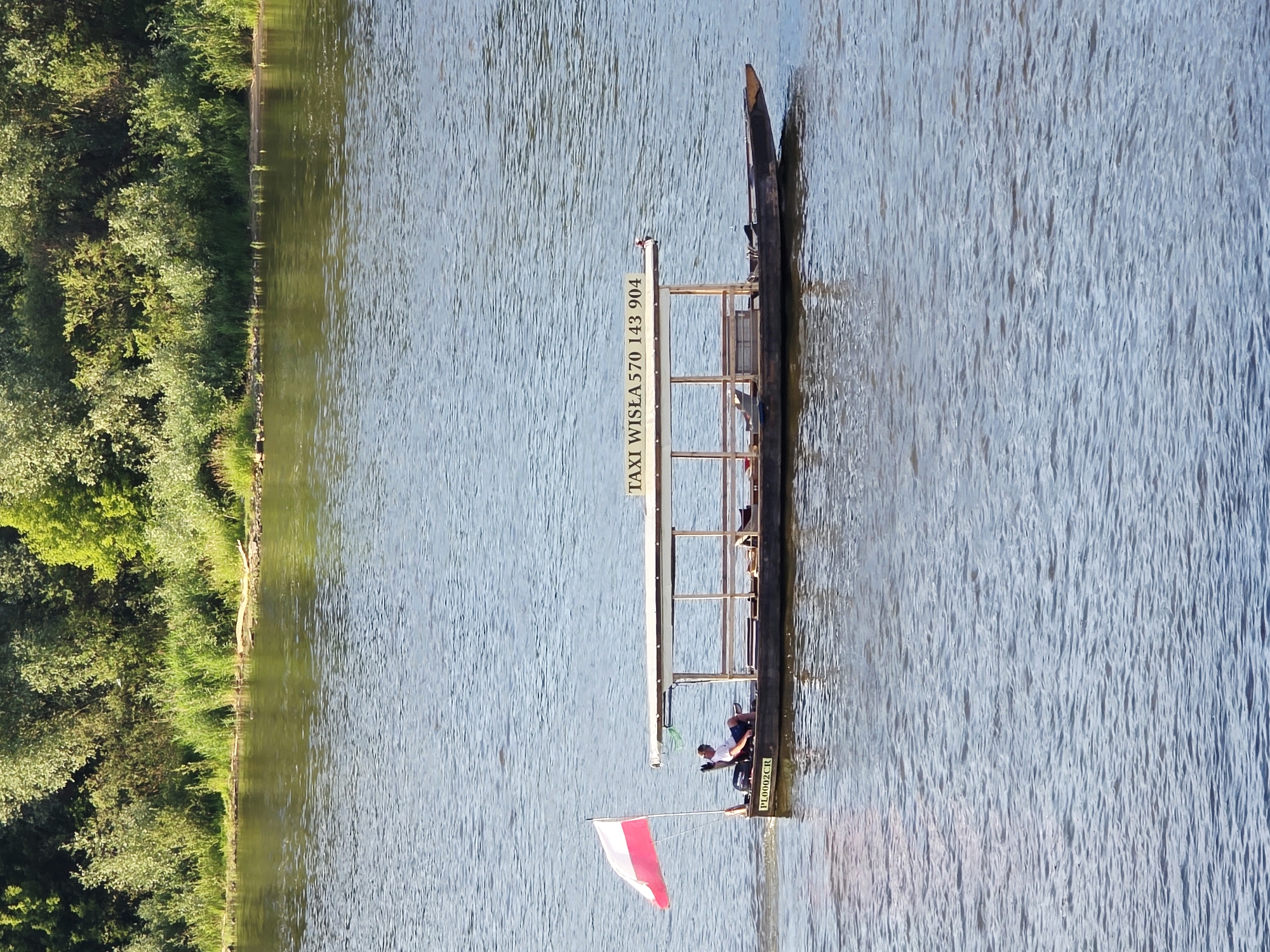 A flat-bottomed boat with a canopy is traveling on a river. The boat is dark brown and has a white canopy supported by wooden posts. The words "TAXI WISŁA 570 143 904" are printed on the canopy in black lettering. A man wearing a white t-shirt and dark shorts is sitting at the helm of the boat, steering it. A Polish flag, red and white, is flying from a pole at the stern of the boat. The boat has the license plate "PL0002CR" on its side. The river is calm and reflects the surrounding green foliage on the far bank. The far bank is densely covered with trees and bushes. The sky is not visible. The scene appears to be on the Vistula River in Warsaw, Poland, given the name "WISŁA" on the boat, which is the Polish name for the Vistula River.