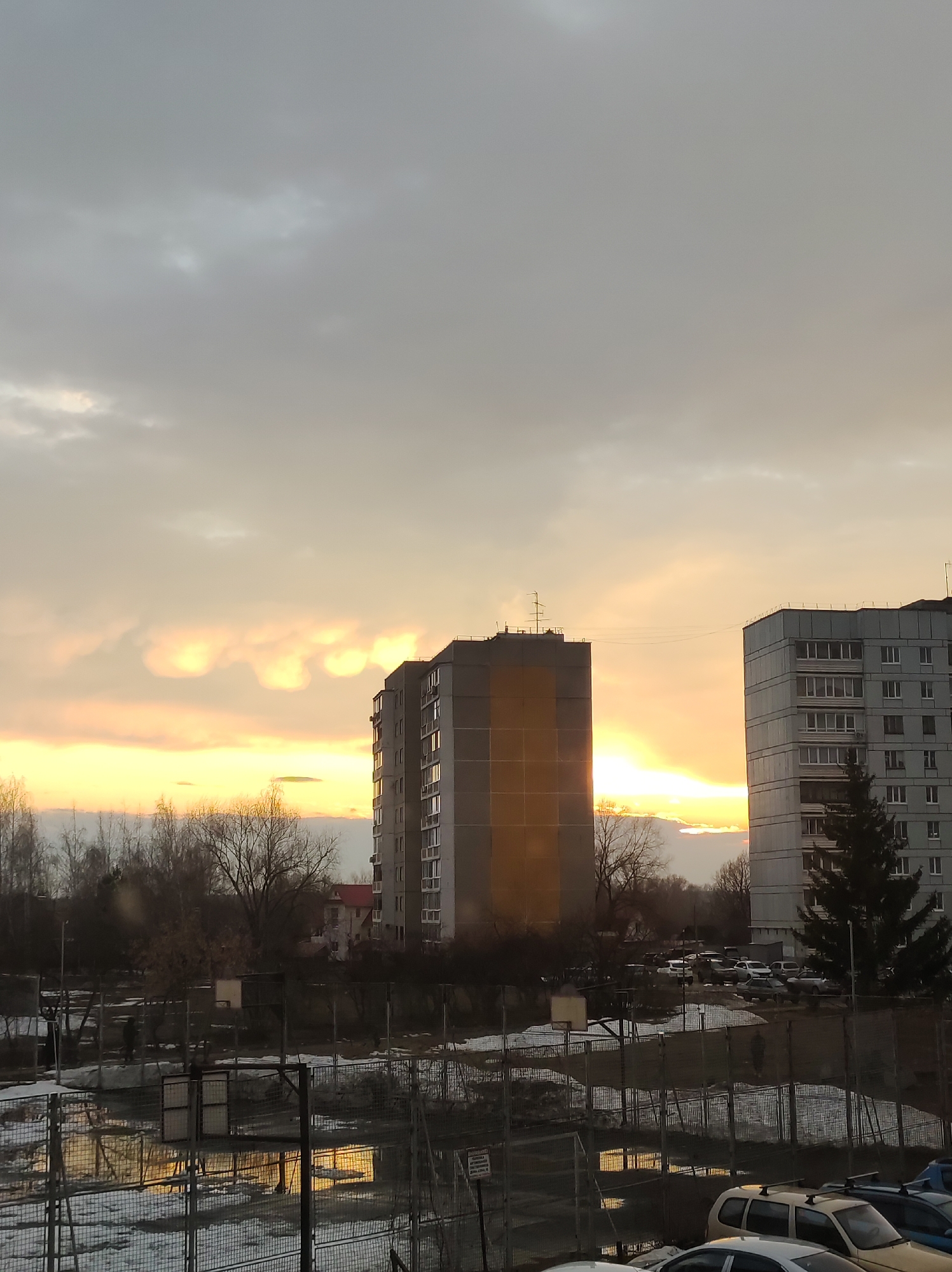 The image captures a cityscape during what appears to be either sunrise or sunset, given the golden hue illuminating the sky. Two multi-story residential buildings dominate the midground; one is predominantly gray with multiple windows, while the other reflects the sunlight, creating a bright, almost monolithic appearance. In the foreground, there's a fenced area that looks like a sports field or a recreational area, with patches of melting snow and puddles reflecting the sky's light. Some cars are parked nearby, suggesting a residential neighborhood. The background features bare trees, suggesting a time of year when the leaves are off the trees, either late fall, winter, or early spring.
