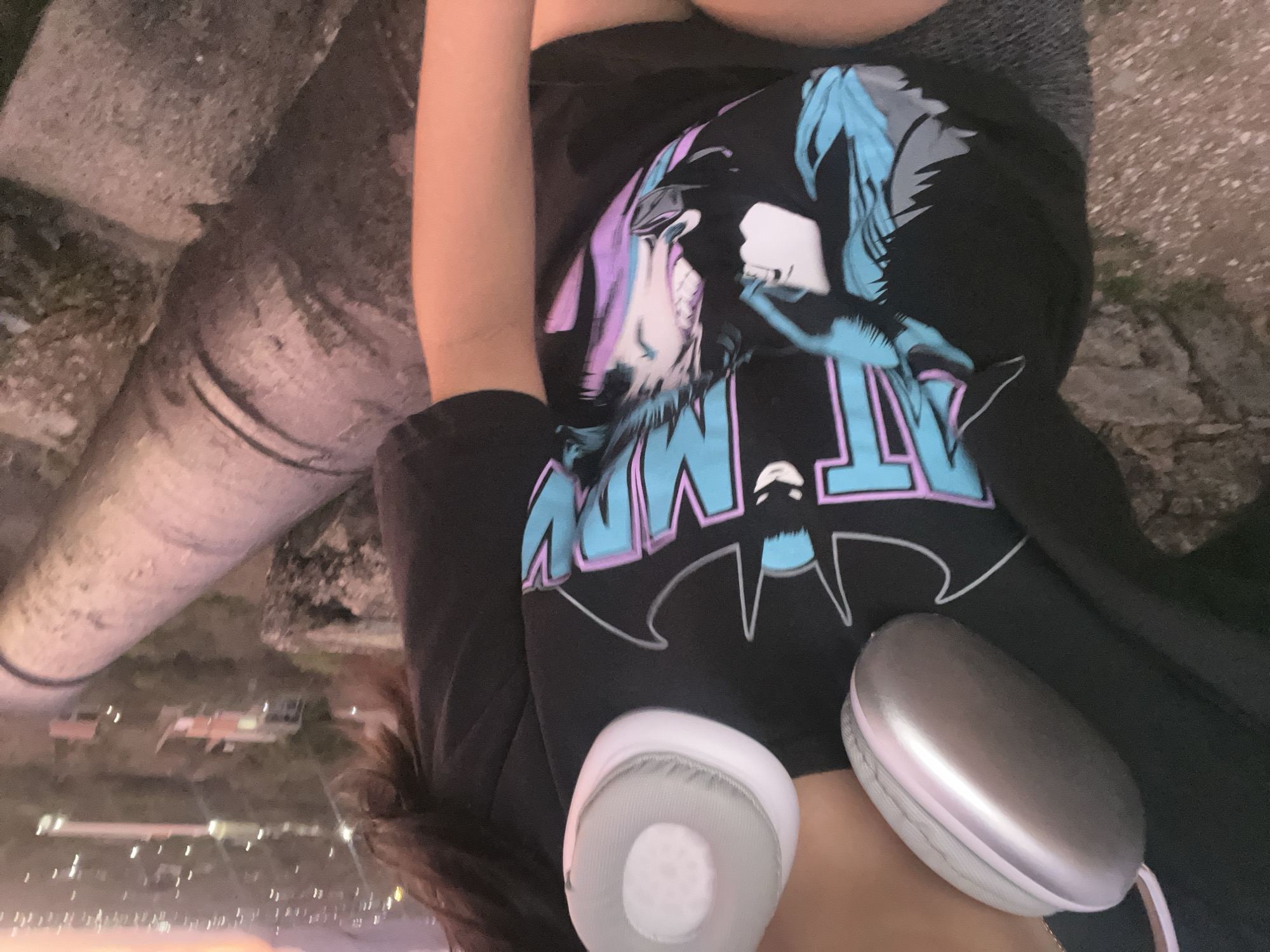 A close-up, mid-shot captures a person wearing a black t-shirt featuring a stylized Batman logo and comic book art. The t-shirt has a Batman logo silhouette and the words "BATMAN" in bold, blue and pink lettering, with comic book panels depicting Batman characters. The person is also wearing silver and white headphones. The background is blurred, but it appears to be an outdoor setting with a cityscape and hills in the distance. A large, weathered cannon barrel is visible to the right of the frame, partially obscuring the cityscape. The lighting suggests it is dusk or dawn. The location is identified as Santa Ana, Venezuela.