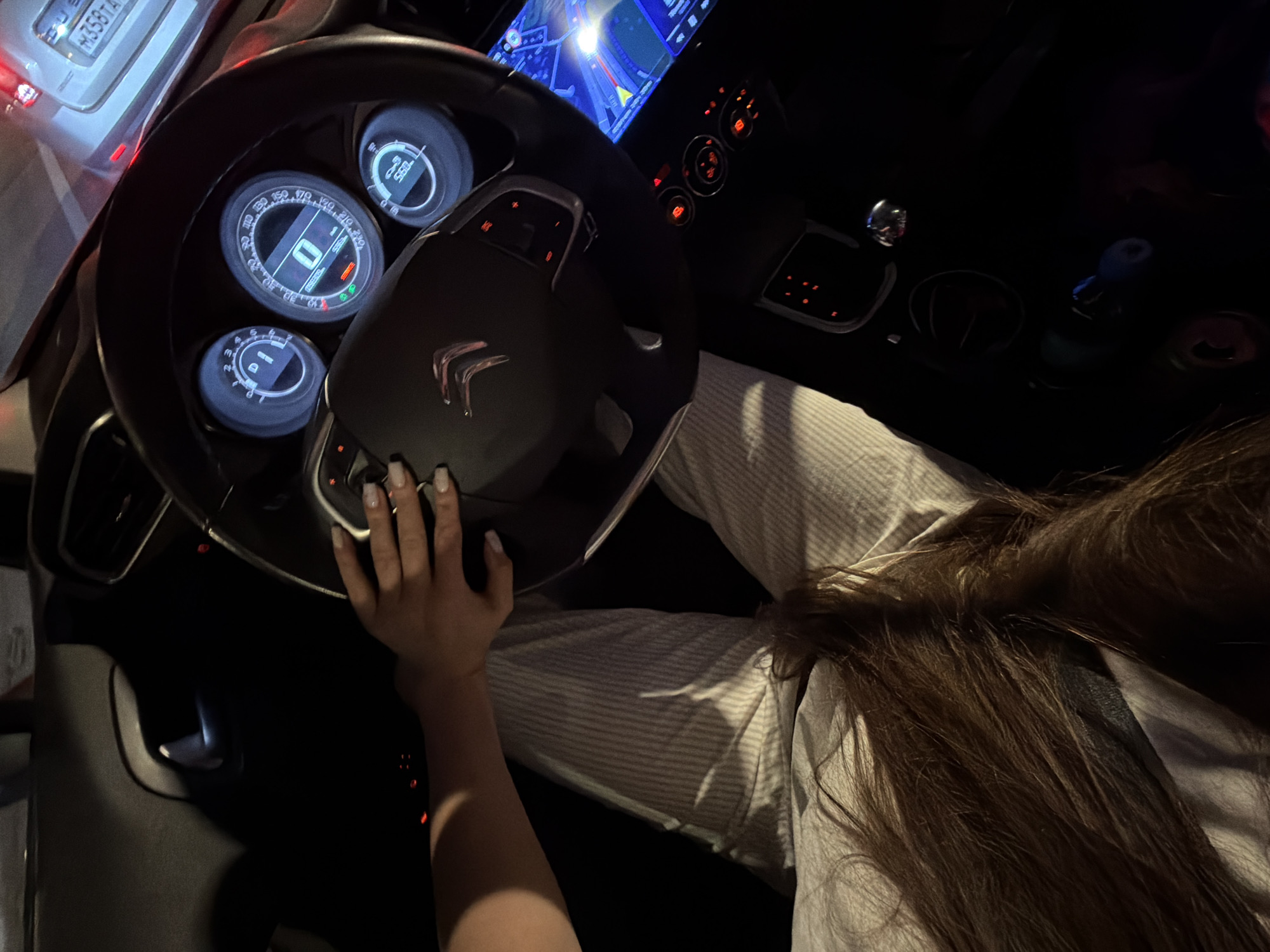 The image shows the interior of a car at night. A person's hands are on the steering wheel, and their legs are visible, wearing striped pants. The dashboard lights are on, displaying the speedometer at 0 km/h. The navigation screen is also illuminated. The exterior of another car is visible through the car's window, with a license plate that appears to be Russian, reading "H358TA". The context suggests the car is stationary in Moscow, Russia.