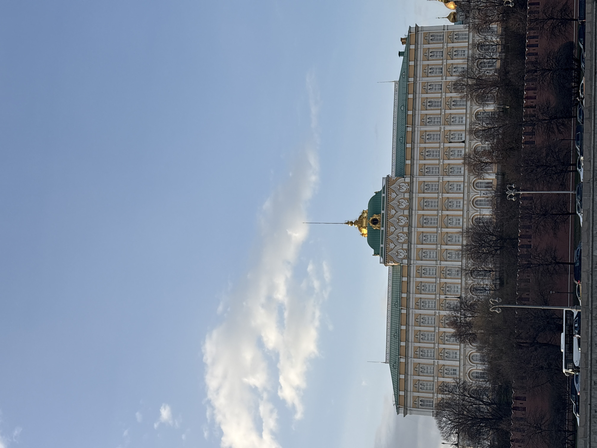 Here's a description of the image:
The image features a portion of the Grand Kremlin Palace in Moscow, Russia. The building is a large, multi-story structure with a pale yellow facade, white trim, and a green roof. The palace has numerous windows arranged in neat rows. Part of a domed roof, topped with a gold spire and cross, is visible. To the right of the palace is a red brick wall, bare trees, and several parked buses. The background shows a clear blue sky with some white clouds. The photo is oriented vertically, with the building positioned to the right of the frame and the sky to the left.