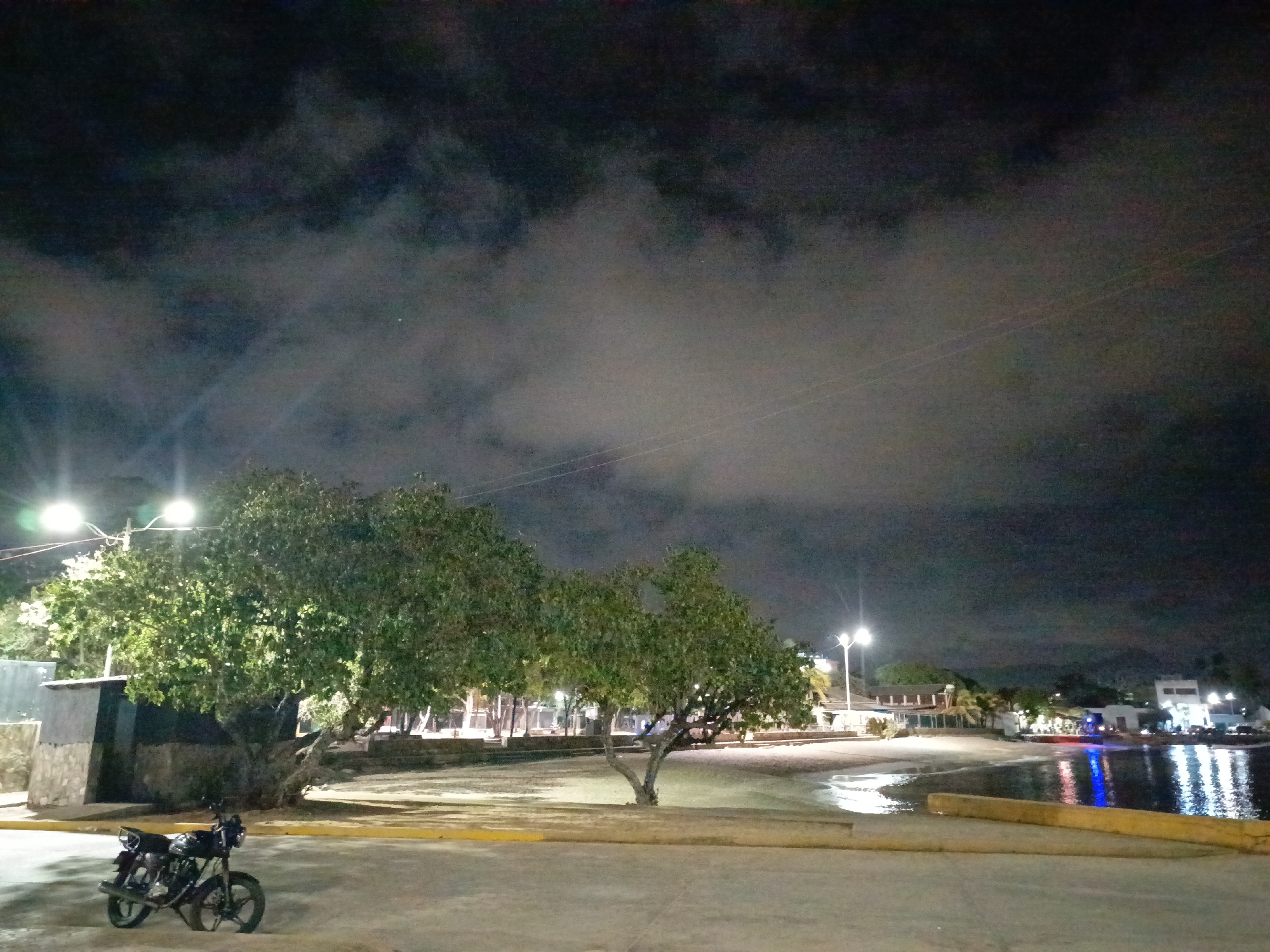 At night in Juan Griego, Venezuela, a motorcycle stands on a concrete pavement in front of a small building and trees. Beyond the trees is a sandy area that meets the water's edge. Lights reflect in the water from buildings in the distance. Above, the sky is cloudy.