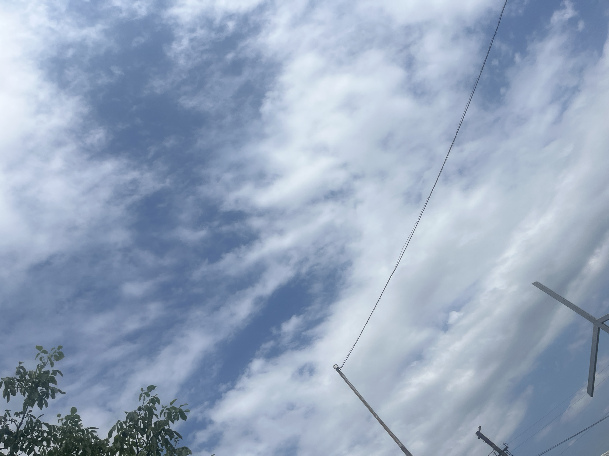 The image captures a bright, cloudy sky with wispy, elongated clouds stretching across a pale blue expanse. In the upper left corner, the dark green leaves of a tree are visible, partially obscuring the sky. A thin, dark wire stretches diagonally across the frame from the lower left to the upper right. To the left of the wire, a tall, thin pole with a ring at its top is angled upwards. Below this pole, a portion of another utility pole with crossbars is visible. In the lower right quadrant, a section of a metallic, angular structure, possibly part of a fence or a sign, is present. The scene appears to be an outdoor view, looking upwards, in the city of Surami, Georgia.