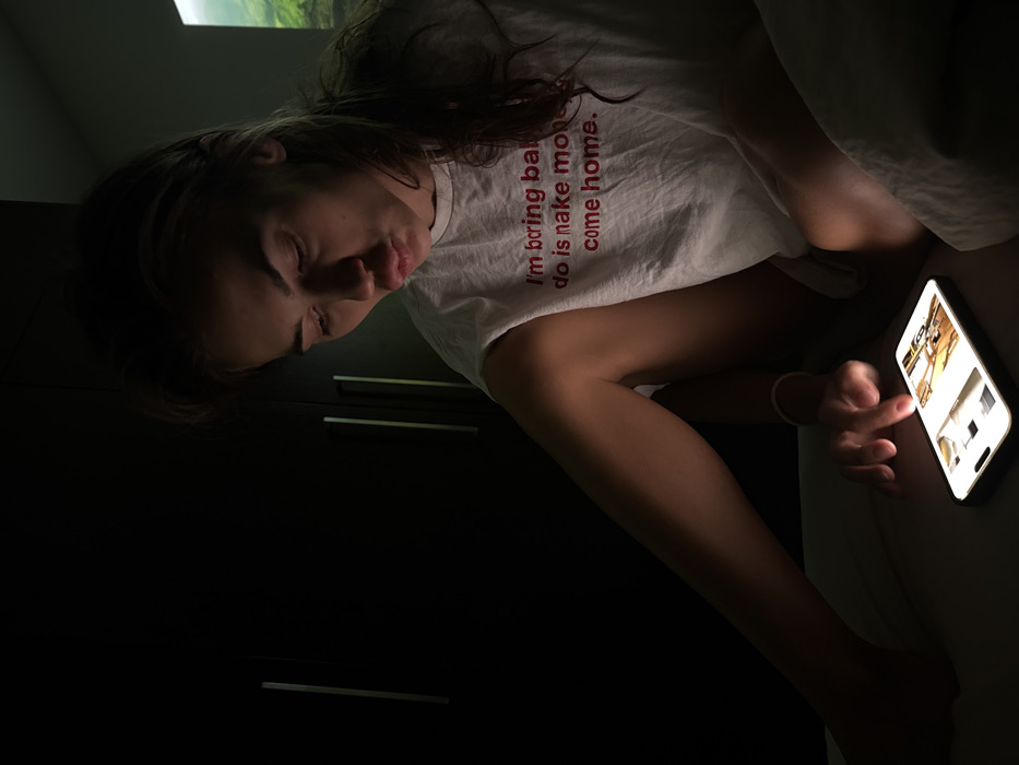 A young woman lies in bed, intently focused on her phone screen. The phone's glow illuminates her face and hand. She wears a white t-shirt with a humorous message printed on it. A dark cabinet stands behind her. The scene is dim, suggesting it's night. The overall impression is of someone engrossed in their device, perhaps late at night.