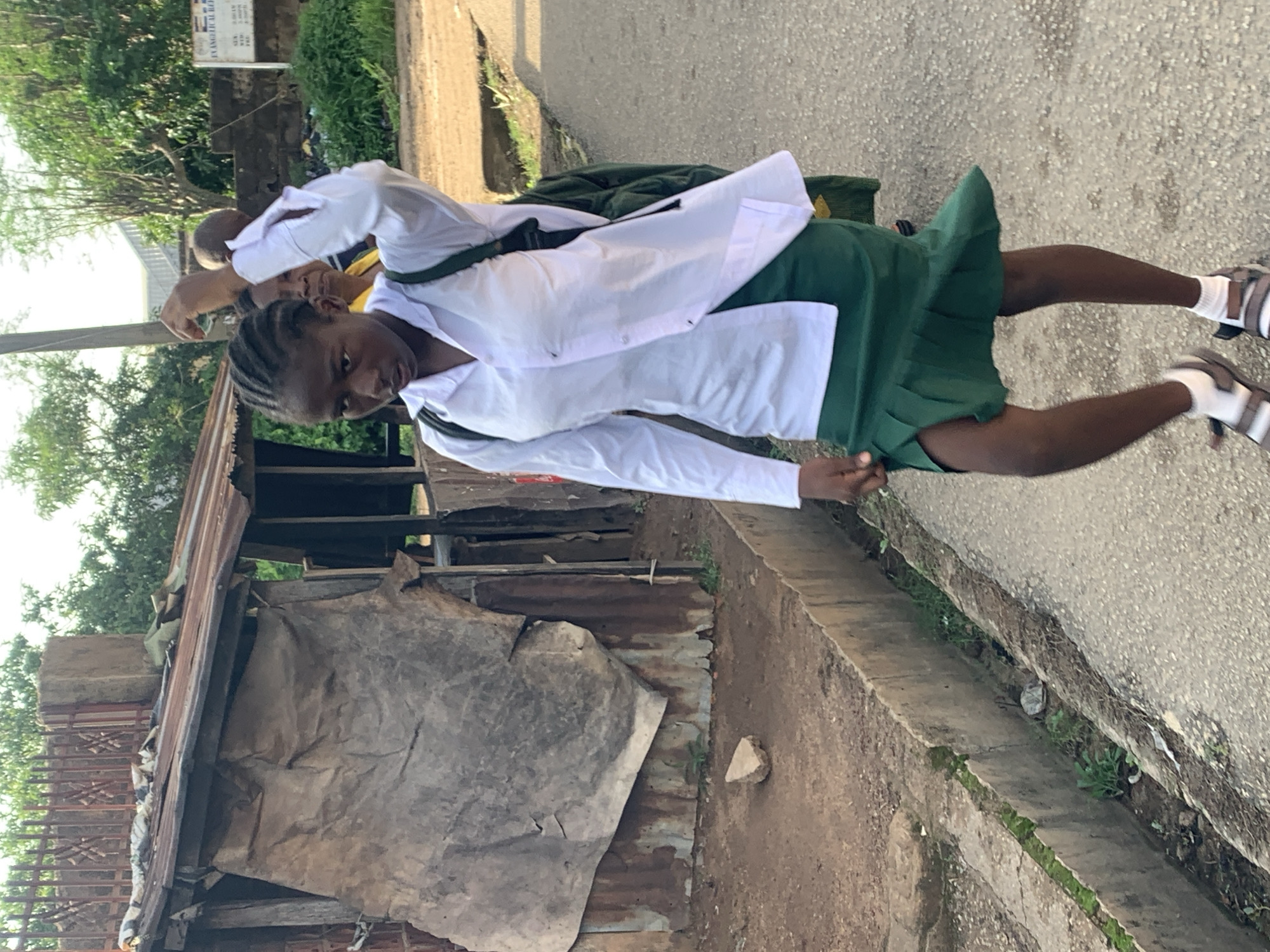 A young Black girl with braided hair and wearing a white collared shirt and a green pleated skirt walks along a paved road in Jos, Nigeria. She has a dark green backpack slung over her left shoulder and is raising her right hand as if to shield her face from the sun or adjust her hair. Her left hand is by her side. She is wearing white socks and grey sandals. Behind her, a smaller Black child, possibly a boy, wearing a yellow shirt, is also walking. To the left of the road is a dirt path and a dilapidated structure with a rusty corrugated metal roof and walls made of various materials, including what appears to be a large, grey tarp. A concrete drain runs along the edge of the road. Trees and foliage are visible in the background, and a sign with text is mounted on a wall. The lighting suggests it is daytime.