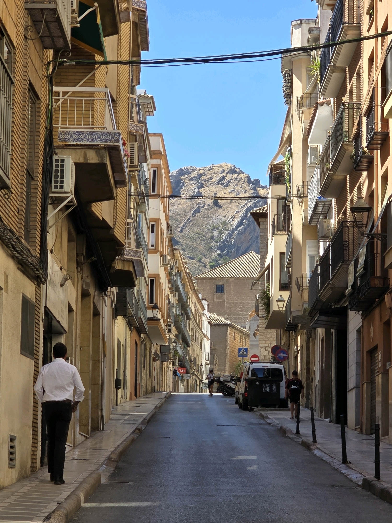 In Jaén, Spain, a man wearing a white collared shirt and black pants walks away from the camera down a narrow, sun-drenched street. The street is lined with multi-story buildings with balconies, some adorned with intricate tile work. The buildings are a mix of warm tones, from ochre to pale yellow and terracotta. Air conditioning units are visible mounted on the facades of several buildings. Power lines crisscross overhead against a clear blue sky. In the background, a rugged, brown mountainside dominates the horizon. Further down the street, a white van is parked, and other individuals are visible in the distance. The street itself is paved with asphalt, with a tiled sidewalk bordering the left side of the road. The overall impression is of a historic European city street on a bright, clear day.