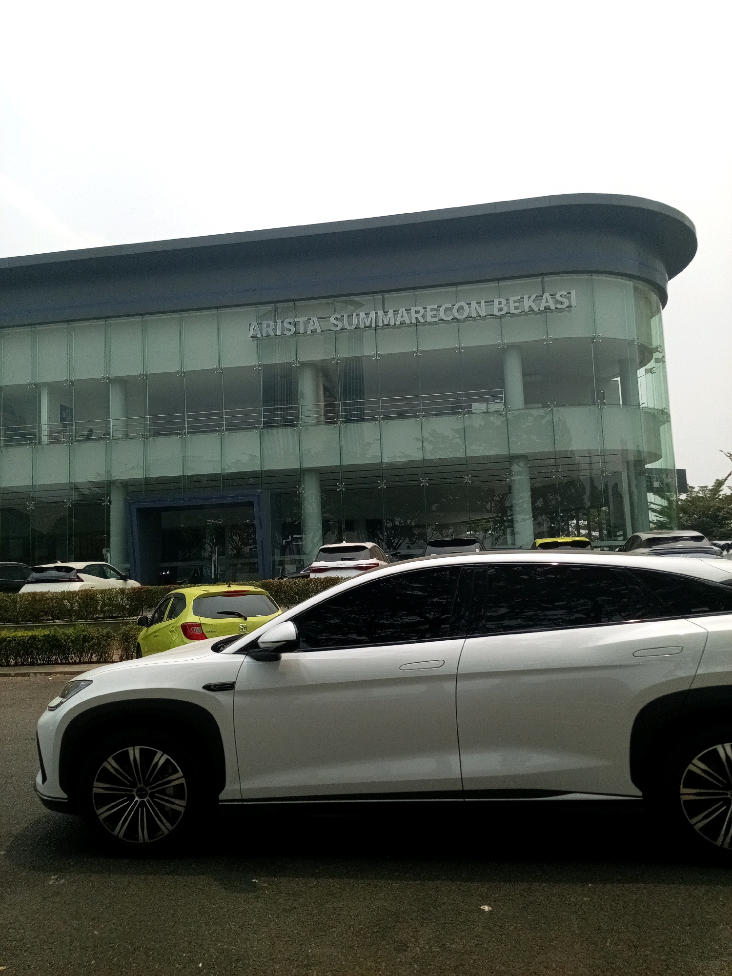 A white SUV is parked in front of a modern glass-fronted building with the text "ARISTA SUMMARECON BEKASI" displayed prominently on its facade. The building appears to be a car dealership, as indicated by the text and the presence of other vehicles visible through the glass, including a yellow car and several other cars parked in front of the building. The SUV is in the foreground, partially obscuring the view of the building and other cars. The environment is an urban setting, likely a street or parking lot in Bekasi, Indonesia, with a slightly overcast sky. The ground is paved with asphalt. There are no people clearly visible in the image.