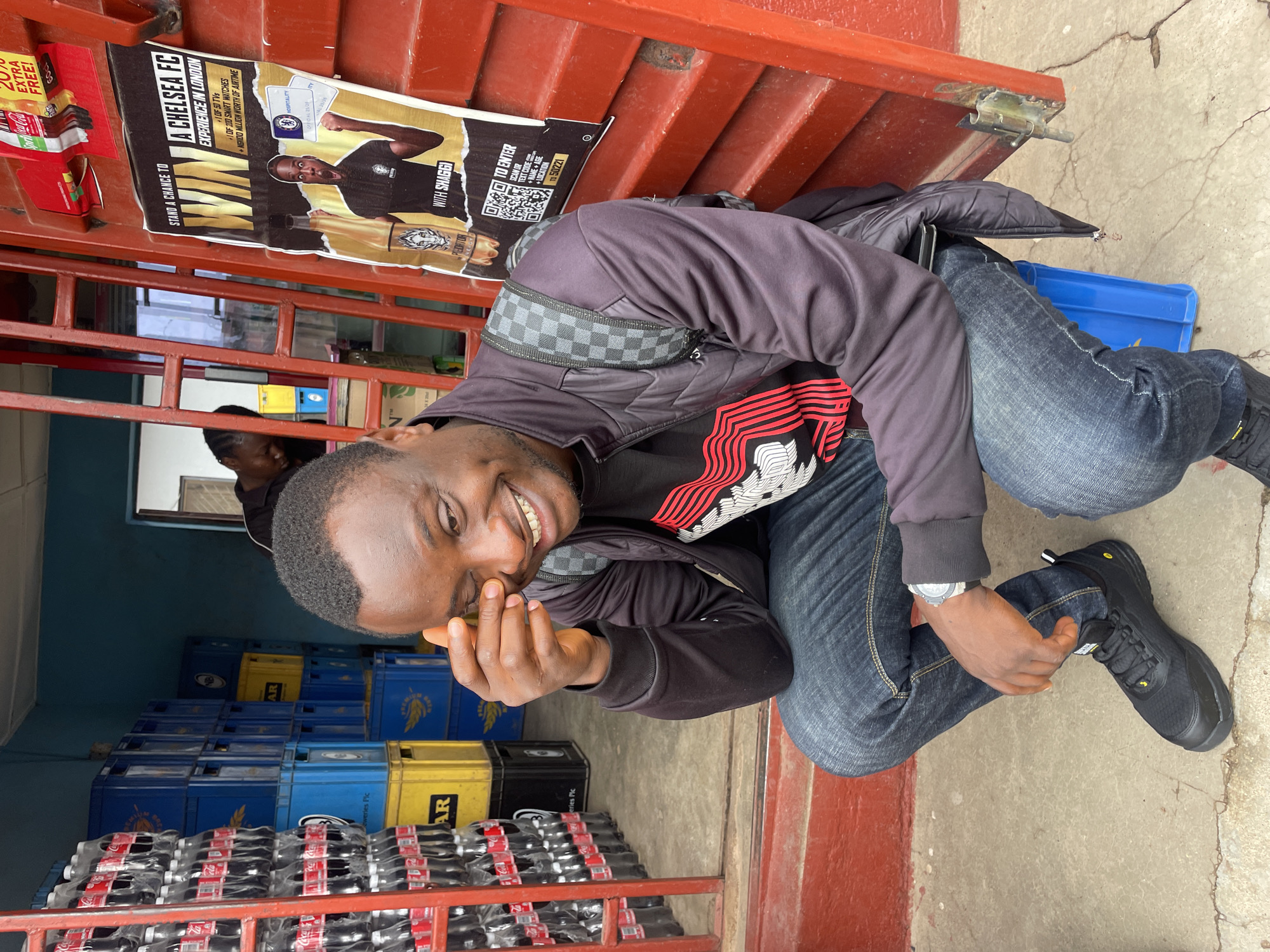 A man is crouched down and smiling at the camera. He is wearing a black jacket with a red and white patterned shirt underneath, blue jeans, and black shoes. He is sitting next to a stack of Coca-Cola bottles and blue and yellow crates. Behind him is a red metal structure with a Chelsea FC advertisement. The ad reads, "A CHELSEA FC EXPERIENCE IN LONDON," and "STAND A CHANCE TO." Based on the text "TEXT CO NAME + LOCATION TO 50221," the image likely depicts a promotional event. Due to the provided information, it's not possible to determine the exact location in the city of Jos, Nigeria, but the setting is likely a retail establishment or event venue.