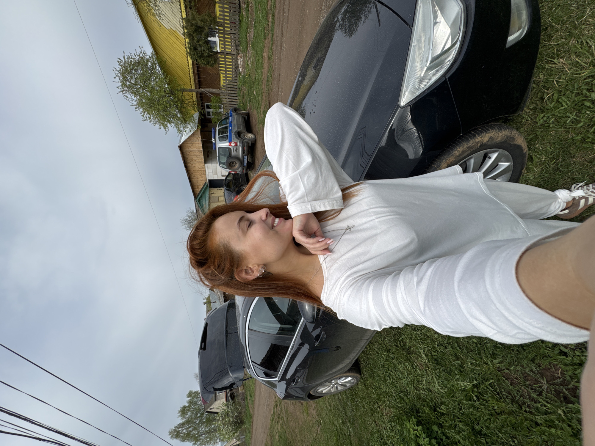 A young woman with red hair, wearing a white shirt, stands outside next to a black car. She is smiling and looking off to the side with her hand near her face. The car is on a grassy area, likely a driveway or roadside. In the background are several buildings, including a yellow one. Another car, possibly a police car, is visible in the distance. The sky is overcast. The location is Chastyye, Russia.
