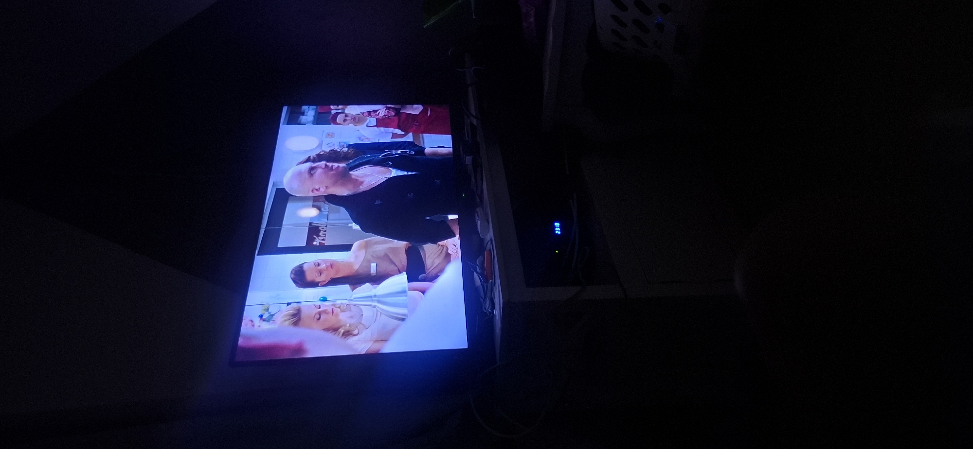 The image shows a television screen displaying a scene with several people. On the left, two women are visible; one has blonde, curly hair and is wearing a light-colored dress. The other woman has dark hair pulled back and is wearing a beige top. In the center of the screen, a bald man with a serious expression is wearing a dark jacket over a white shirt and a silver necklace. To his right, another person is wearing a red apron, suggesting a food service or butcher shop setting. The background of the television screen appears to be an interior with white walls and some shelving.
Beneath the television, a white media console is visible. A blue digital display on the console shows "23:18". Various cables and electronic devices are present on and around the console. The overall environment is dark, with the television screen being the primary light source.
This scene is not located in Yerocham, Israel.