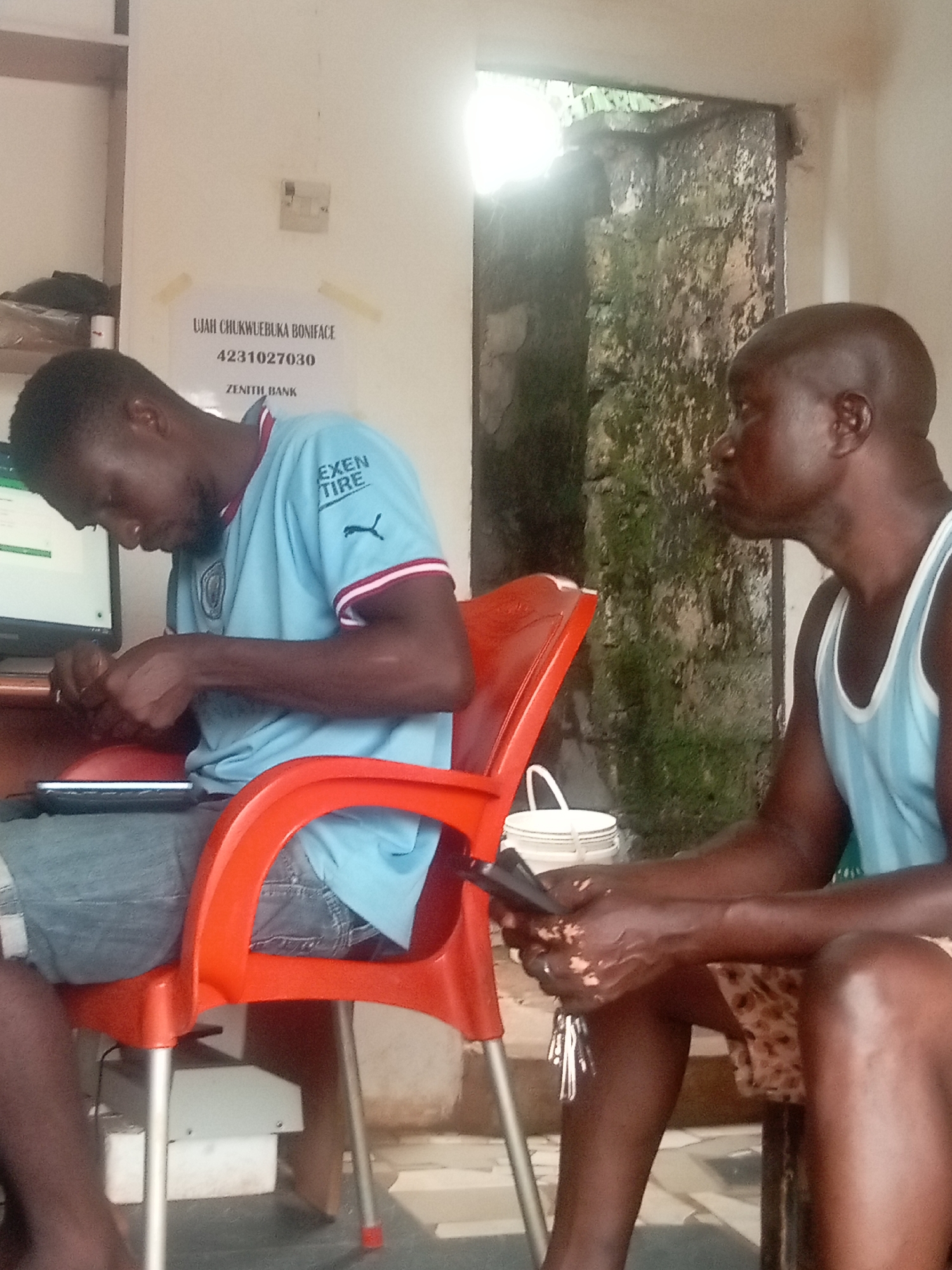 In Ogidi, Nigeria, two dark-skinned men are indoors. The first man is seated on an orange plastic chair, wearing a light blue t-shirt, and light blue jeans. He is looking down at a tablet device he is holding. The second man is wearing a light blue tank top and patterned shorts. He is seated on a chair, and is holding a mobile phone. A white bucket is visible near the doorway in the background. A sign affixed to the wall reads "Ujah Chukwuebuka Boniface" followed by the account number "4231027030", and the bank name "Zenith Bank". A computer monitor sits on a table to the left. The wall near the doorway is covered with a rough, textured substance.