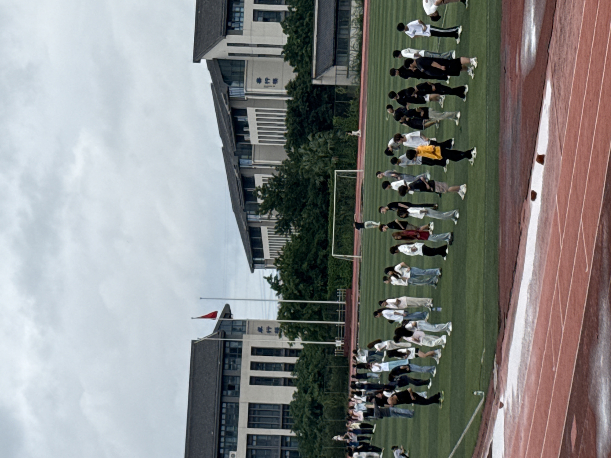 The image shows an outdoor scene on a cloudy day. In the background, two multi-story buildings with dark roofs are visible, possibly part of an educational institution. In front of the buildings, there are trees and a red flag on a flagpole. A green field, likely a sports field, occupies the middle ground, with a goalpost on the right. Numerous people, appearing to be students, are scattered across the field, many standing in lines or small groups. Some are wearing casual clothing like t-shirts and jeans, while others are in athletic attire. A running track, colored red and with white lines, is in the foreground. The track surface appears wet in some areas, possibly from rain. The overall impression is of a school or university campus in Jiangjin, China, with students engaged in an outdoor activity or assembly.