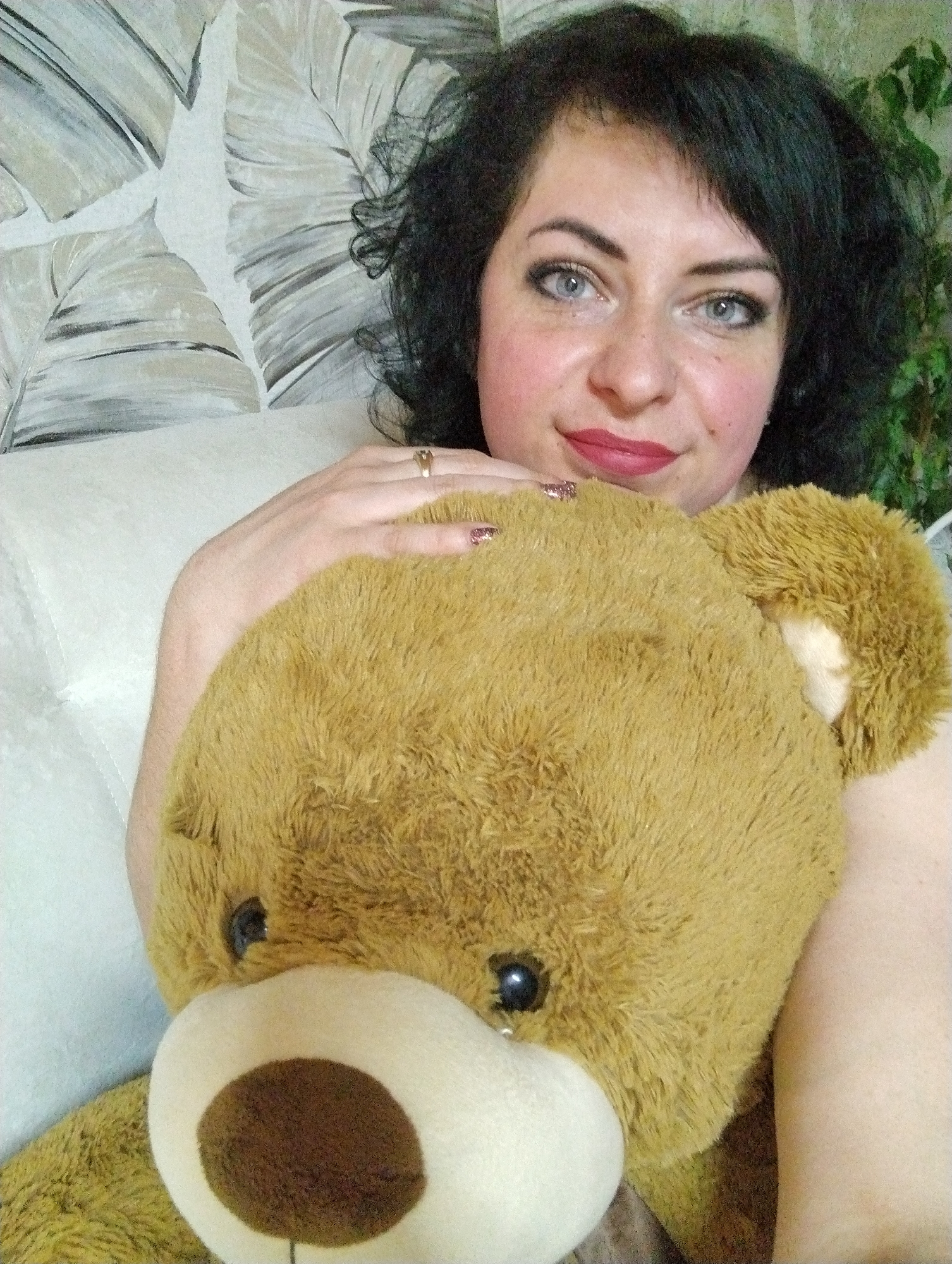 A woman with dark curly hair and blue eyes is holding a tan teddy bear. She has fair skin, wearing red lipstick, and a gold ring on her left ring finger. The teddy bear has a white snout and brown nose. The background consists of a white sofa and a wall with a palm leaf pattern. Green foliage is visible in the upper right corner. The location is not discernible from the image.