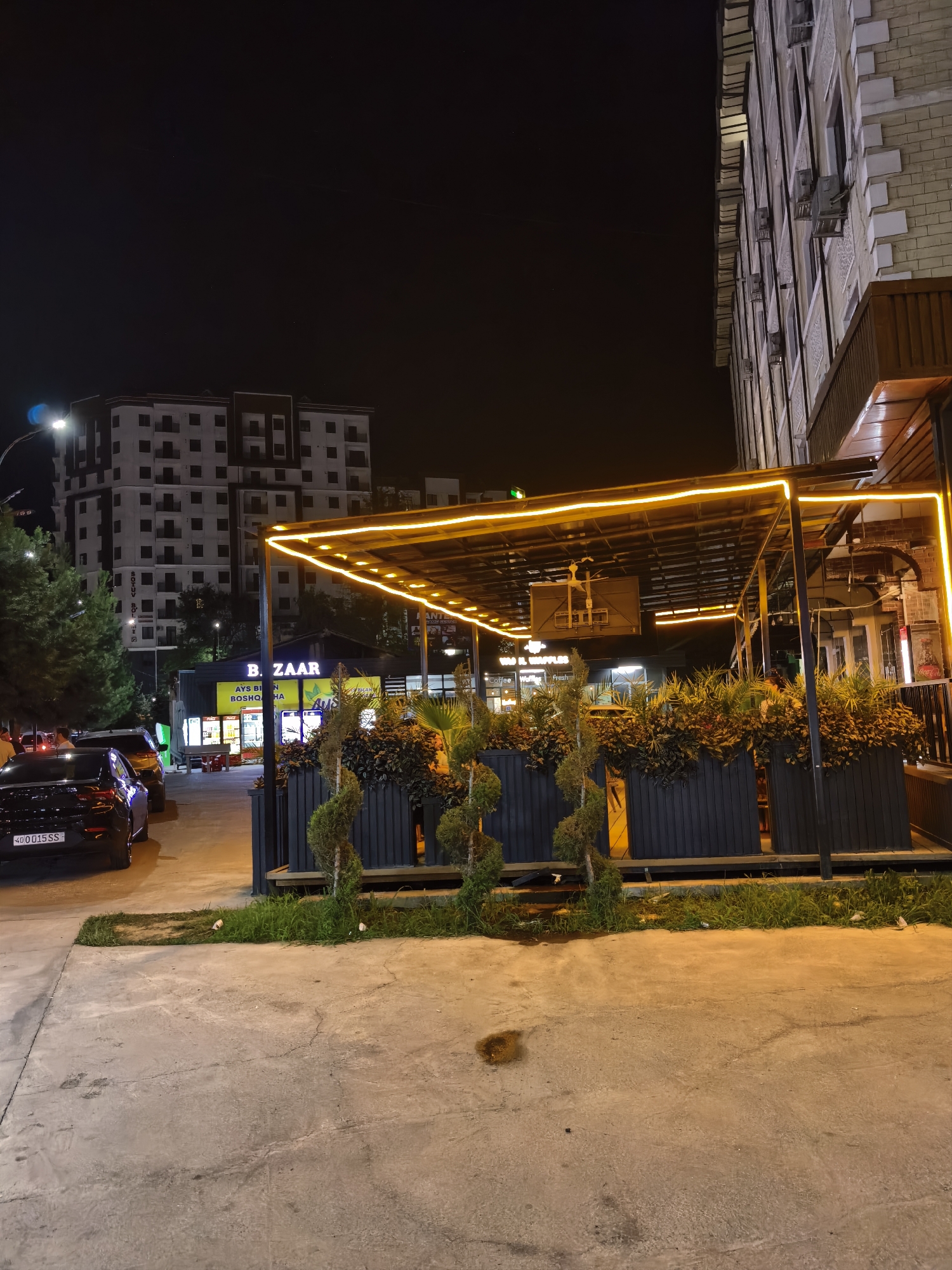The image depicts a nighttime scene in Fergana City, Uzbekistan, focusing on a commercial establishment with an outdoor seating area. The establishment appears to be a restaurant or cafe, with a covered patio adorned with bright yellow neon lighting. A dark-colored sedan is parked on the left side of the image, its license plate visible, showing "40 0015 SS". In the background, a multi-story building with numerous windows is illuminated. Adjacent to the main establishment, a smaller storefront with the word "BAZAAR" prominently displayed is visible. The ground is paved with concrete, and there are planters containing greenery and decorative topiaries in front of the seating area. The overall atmosphere suggests a lively urban commercial district at night.