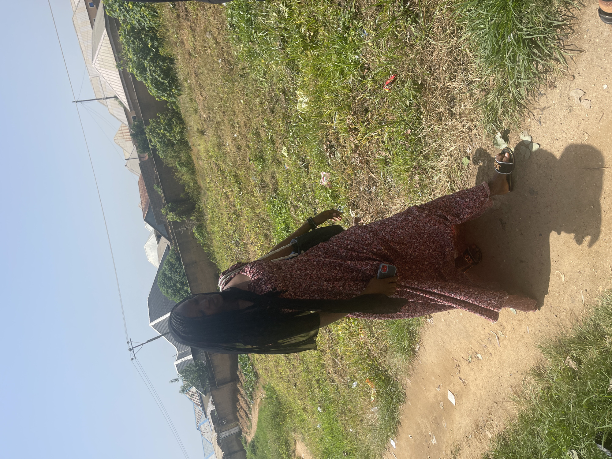A full-body outdoor shot shows a young woman standing on a dirt path, bathed in bright sunlight, suggesting a warm, clear day. She is positioned in the center of the frame, facing slightly away from the camera, with her hair braided and falling down her back. She wears a long, floral-patterned dress with a deep V-neck and long, flowing sleeves, and black sandals. In her right hand, she holds a smartphone with a red case. The background reveals a grassy area with some scattered debris, and a line of buildings with white roofs and gray walls are visible on the left side of the image. The overall setting appears to be a residential area in Rafin Par, Nigeria. The lighting and shadows suggest it is likely midday. The woman's posture and relaxed stance indicate a casual and possibly pleasant moment outdoors. There is no visible text in the image.