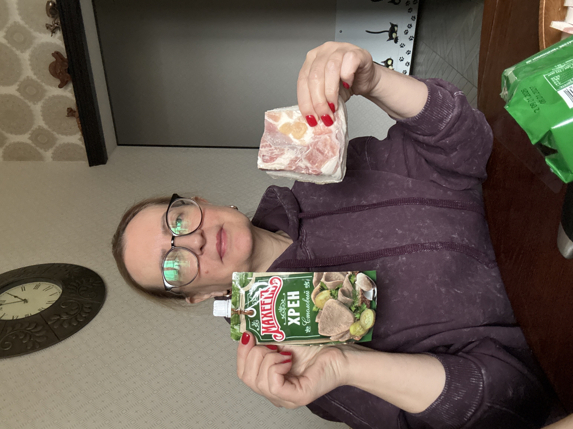 A woman is depicted indoors, likely within a home setting in Krapivka, Russia. She is wearing glasses and a dark purple long-sleeved top. The image, which appears to be a selfie, shows her presenting two food items.
In her right hand, she holds up a rectangular block of what seems to be frozen meat or lard, characterized by layers of pinkish meat and white fat, with a small yellowish portion visible. Her fingernails are painted bright red. In her left hand, she displays a green flexible pouch of "МАХЕЕВ ХРЕН Столовый" (Makheev Table Horseradish). The packaging features an illustration of sliced meat, possibly tongue, served with pickled cucumbers and dill.
The background consists of a light-colored wall covered in wallpaper with a subtle, repeating geometric pattern. To her left, a partially visible, ornate dark-framed wall clock with Roman numerals indicates a time near the top of the hour. Above her right shoulder, a dark-framed item, possibly a screen or mirror, is visible, with small decorative figurines positioned on a shelf above it. To her right, a dark wooden table holds a green package with Russian text, including dates "2.09.01.2008" and "08.01.2027", along with words like "ПОЛОЦКИИ" (Polotskiy) and "ЭКСТР" (Extra). A decorative element featuring black cat silhouettes and paw prints adorns the wall in the upper right. The woman maintains a neutral expression, and the scene suggests a domestic activity such as preparing or presenting food.