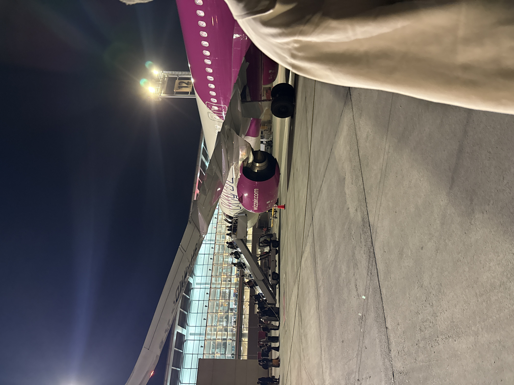 The image depicts an airplane parked on an airport tarmac at night. The main subject is a Wizz Air aircraft, distinctively colored in pink/purple on its upper fuselage and engine cowlings, contrasted with a white lower fuselage and wings.
Numerous people, likely passengers, are seen on an airstair that leads to the aircraft's door, suggesting an active boarding or disembarking process. More individuals are visible on the tarmac near the base of the airstair and further back, appearing to queue or wait. In the background, a modern airport terminal building with large, illuminated glass windows reflects the interior lights. The foreground features a partial view of a person's light-colored clothing, indicating the photograph was taken from a close, personal perspective next to the aircraft.
The activity taking place is the movement of passengers to or from the aircraft. The scene is set at night, characterized by the dark sky and the strong reliance on artificial lighting from airport structures, the plane itself, and the terminal building, which casts bright glows and some lens flares. The tarmac is visible, with typical ground markings. The weather appears clear. This bustling night-time airport scene is set in Yerevan, Armenia.
Visible text includes "wizzair.com" clearly displayed on the engine cowling. Part of the airline's name, "WIZZ," is also discernible on the side of the fuselage near the tail. Additionally, the number "21" is visible on a floodlight fixture or a piece of ground equipment near the plane.