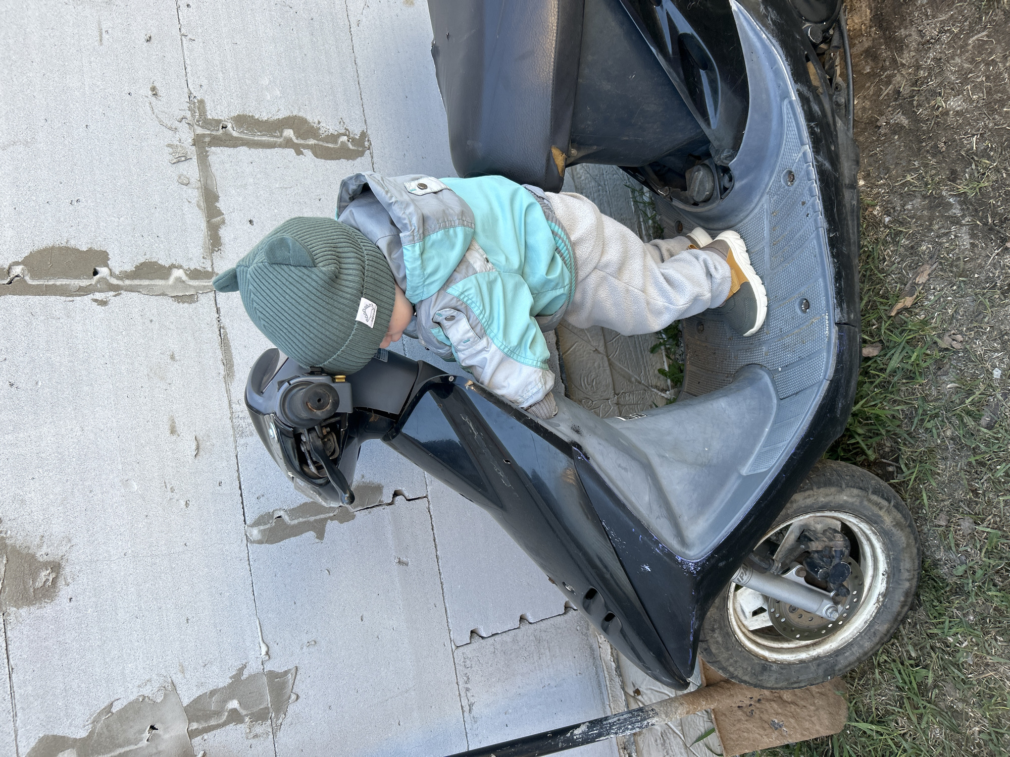 A young child wearing a green hat, a light blue and grey jacket, and grey pants lies prone on a black and grey scooter. The child is positioned on the footrest and seat of the scooter, with their head near the handlebars. The scooter is parked on a surface composed of light grey concrete blocks and patches of dirt with grass. The location is likely outside, given the presence of the grass and dirt. The surrounding area is blurry.