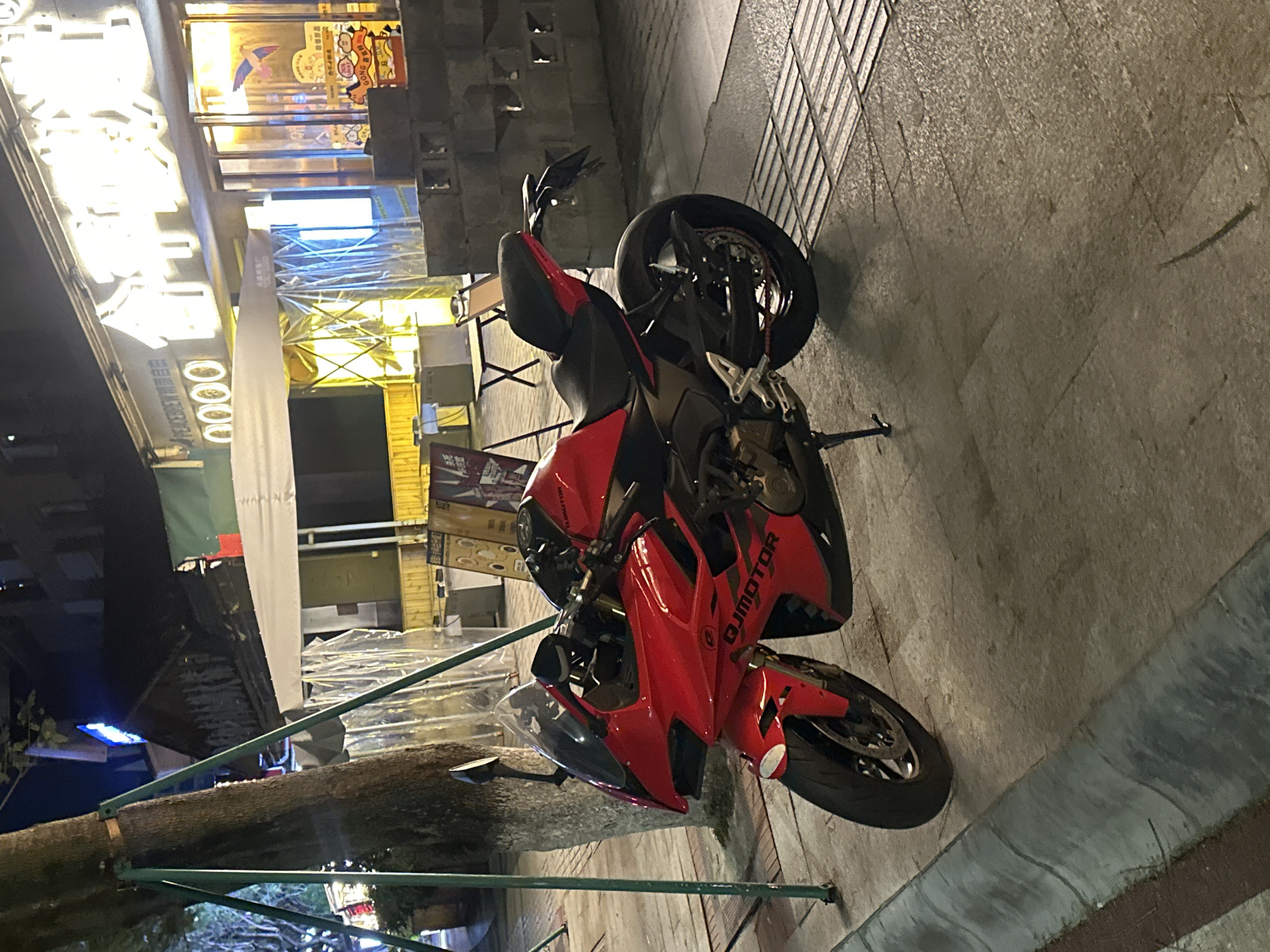 A red and black sportbike motorcycle is prominently parked on its kickstand on a paved sidewalk at night. The motorcycle is the central focus, facing generally towards the left of the frame. Its fairings are primarily red with black accents, and the brand "QJMOTOR" is visible in white lettering on the side fairing.
The setting appears to be an urban street or commercial area in the Chongqing High-tech Zone, China, as suggested by the bright, illuminated storefronts in the background and the visible Chinese characters. The time of day is night, indicated by the artificial lighting from the streetlights and various signs. The ground is paved with square, textured tiles.
In the background, on the left, are brightly lit storefronts. One sign, largely overexposed, clearly shows "PEACEBREW" followed by Chinese characters that appear to translate to "Craft Beer". Other Chinese characters are visible on various signs, though most are too bright or distant to decipher, with one displaying "GONG..." and another "0000". Some temporary structures or scaffolding covered in plastic sheeting are also visible behind the motorcycle. A thick tree trunk, supported by green metal bars, is situated in the foreground on the left, partially obscuring the view of the street beyond. There are no people visible in the image.