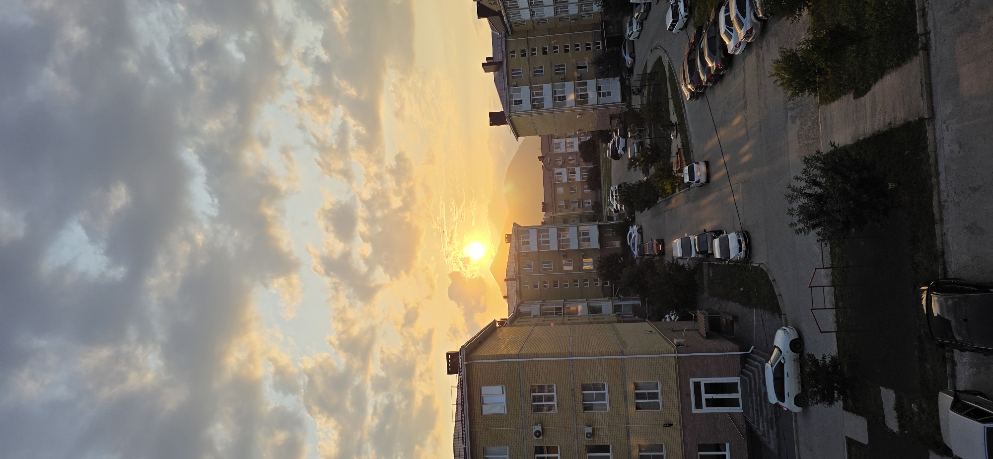 A vertical, wide-angle, eye-level outdoor photograph captures an urban landscape in Inozemtsevo, Russia, at sunset. The sky is filled with a dramatic display of clouds, with the bright, setting sun positioned behind a mountain range. The sun's golden rays illuminate the clouds and cast long shadows across the scene. In the foreground, multi-story apartment buildings with yellow facades and red-tiled roofs dominate the view. A street runs between the buildings, lined with parked cars. The street is paved, and there are patches of grass and trees visible, some with children's playground equipment. The overall impression is one of a typical residential area at dusk.