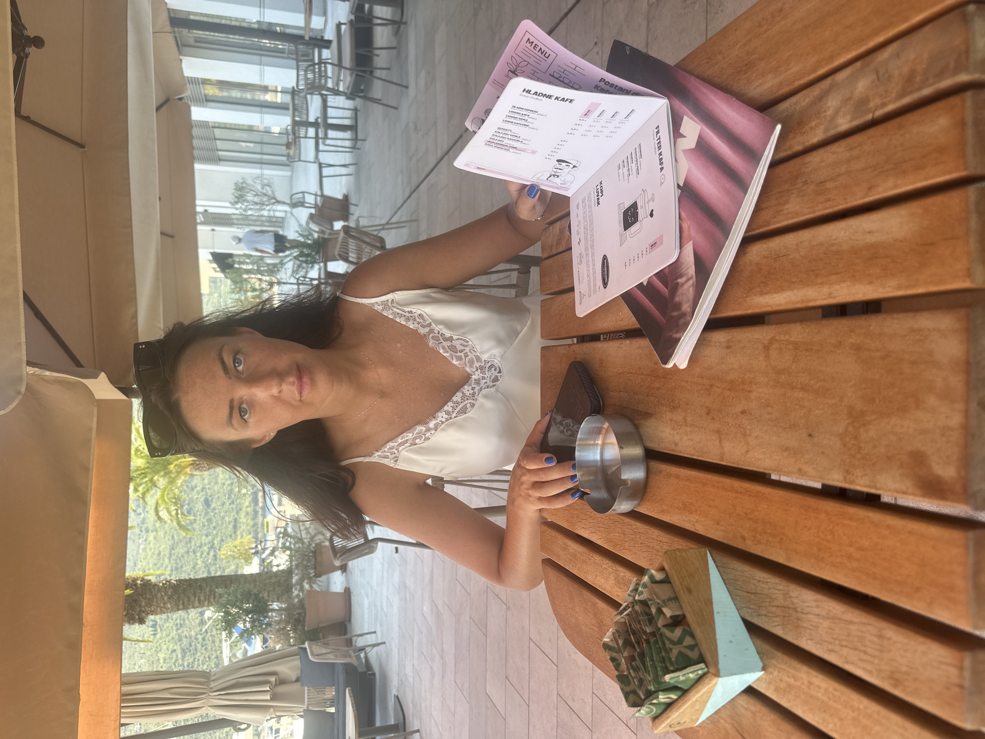 A woman with dark hair and blue eyes, wearing a white satin slip dress with lace trim and black sunglasses perched on her head, sits at a wooden table outdoors. She is looking directly at the camera. In front of her is a mobile phone, an ashtray, and a small wooden container with packets of sugar. She is holding an open menu with both hands, her left hand is resting on the table, and her right hand is holding the menu open. The menu has pink and white pages with text and illustrations, including a drawing of a coffee cup. The table is made of light-colored wooden planks. In the background, there are palm trees, a building with large windows, outdoor seating with tables and chairs, and a beach umbrella. The scene appears to be a cafe or restaurant in Kumbor, Montenegro, with views of vegetation and modern architecture.