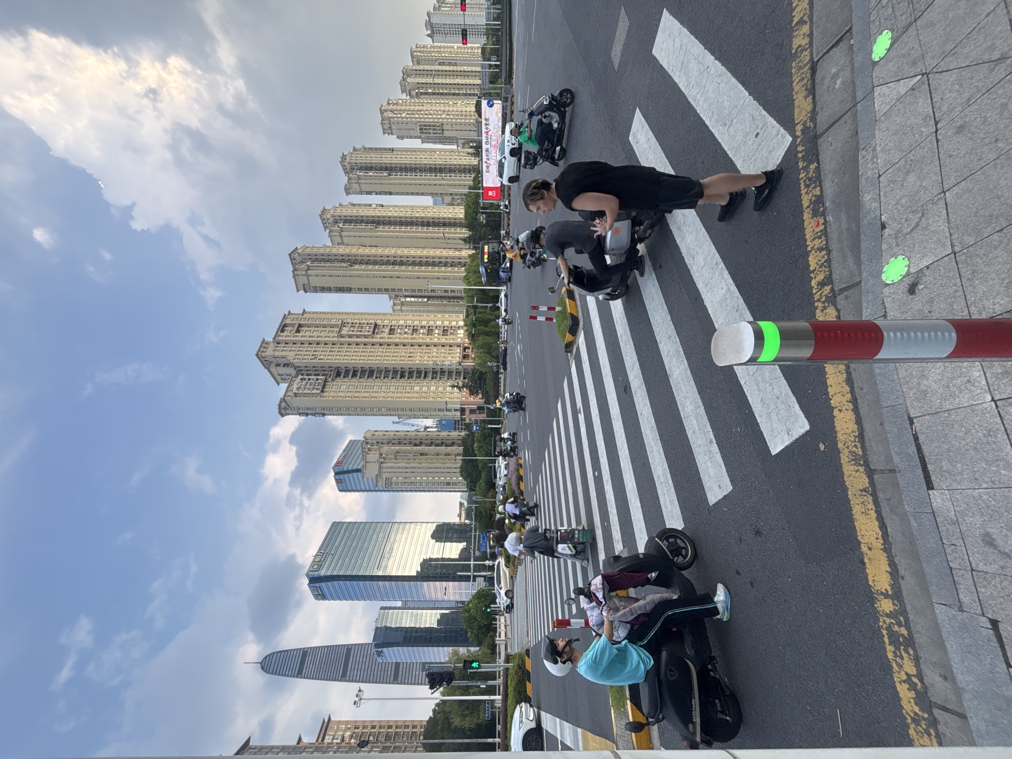 The image depicts a street scene in Suzhou Industrial Park, China, featuring a busy intersection with a pedestrian crossing. Several individuals are seen operating electric scooters, with some stopped at the traffic light and others proceeding through the intersection. The background is dominated by modern high-rise buildings and a clear sky with scattered clouds. A man wearing a black t-shirt and shorts stands in the foreground on the pedestrian crossing, facing away from the camera. Another person on a scooter is visible to his left, wearing a blue t-shirt and helmet. A red and white striped bollard with a green light is positioned on the side of the road. The overall impression is of a dynamic urban environment with active transportation.