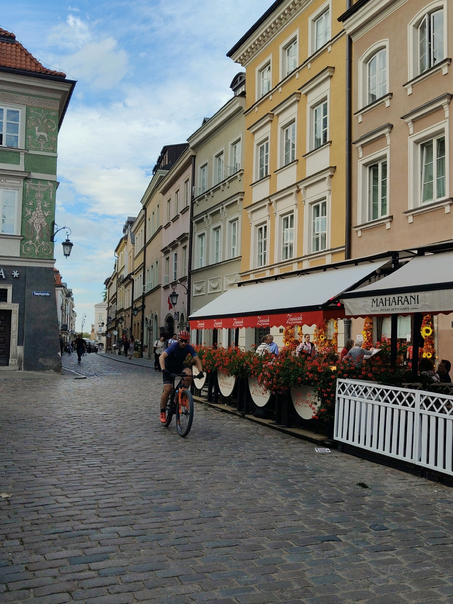 The image depicts a street scene in Warsaw, Poland, likely in the Old Town area, characterized by cobblestone paving and historic architecture. A man wearing a red helmet and cycling attire is riding a bicycle down the street, which is lined with multi-story buildings. The buildings feature various colors and architectural styles, common in European cities. On the right side of the street, there is an outdoor seating area for a restaurant or cafe, adorned with red flowers and white awnings. One awning displays the name "MAHARANI INDIAN RESTAURANT." People are visible at tables in the outdoor seating area and walking further down the street. The sky is partly cloudy. The overall atmosphere suggests a pleasant day in a European urban environment.
