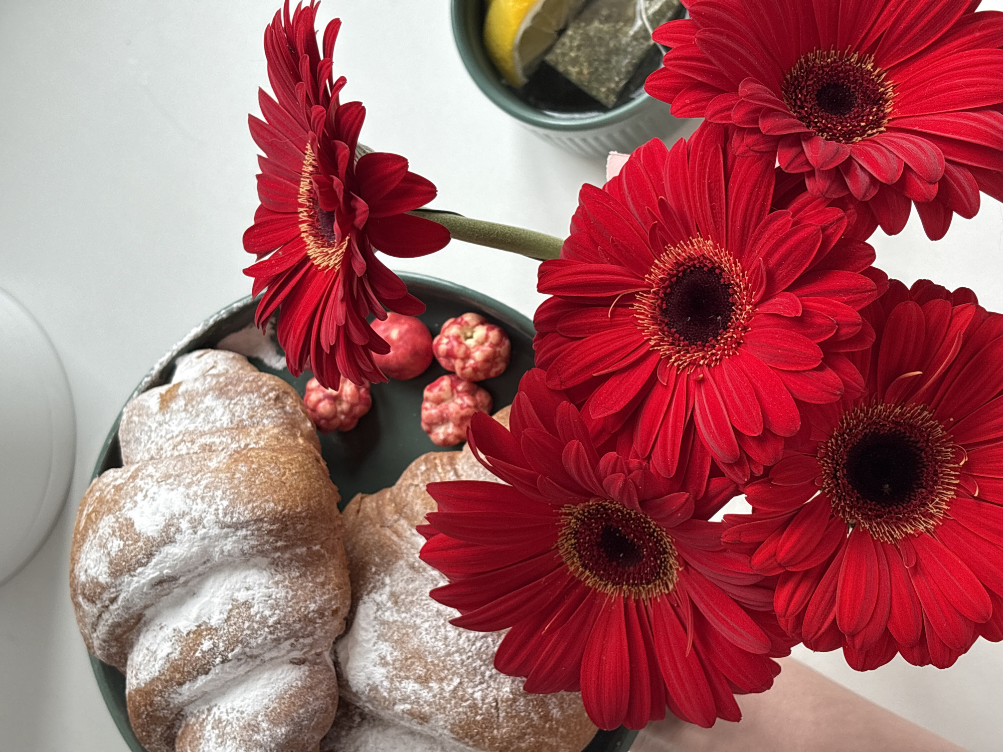 The image depicts a breakfast or brunch scene. A plate with croissants dusted with powdered sugar and small round candies is positioned next to a cup containing a lemon slice and a tea bag. Red gerbera daisies are arranged around the plate and cup. The background is a plain white surface. There is no indication that this image was taken in Chaikovsky, Russia.