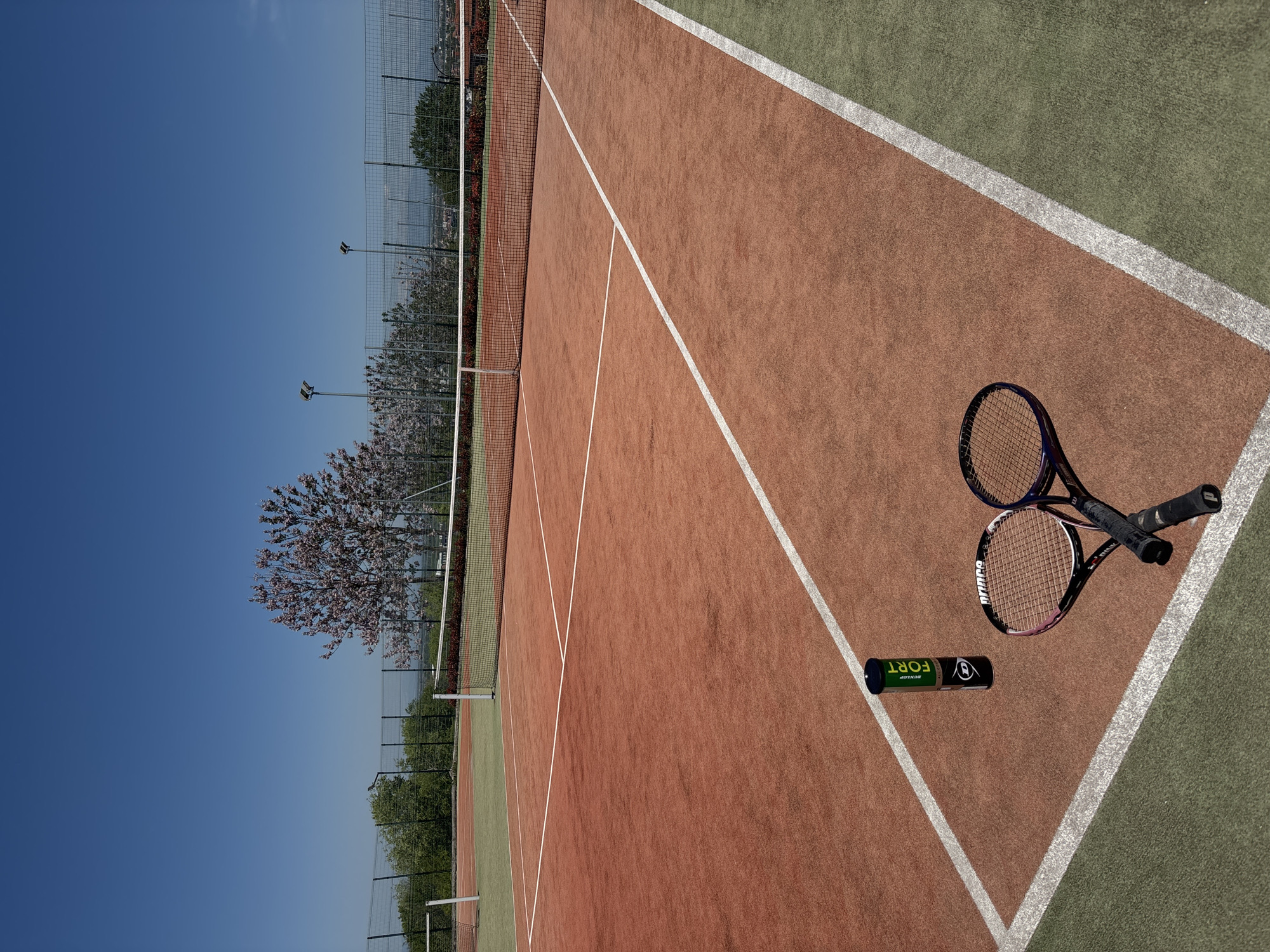 Here is a detailed description of the image:
The image depicts a tennis court under a clear blue sky. The court is a mix of red clay and green hard court surfaces, delineated by white lines. A net stretches across the center of the court. Two tennis rackets, with black grips, are lying on the ground near a can of Dunlop Fort tennis balls. In the background, trees and a fence are visible, suggesting the court is located in a park or recreational area. The location is Unknown, Unknown.